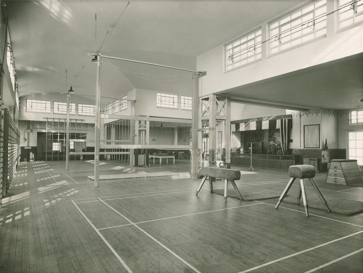 A black and white photo of an interior hall with gymnasium equipment including balance beams and horses