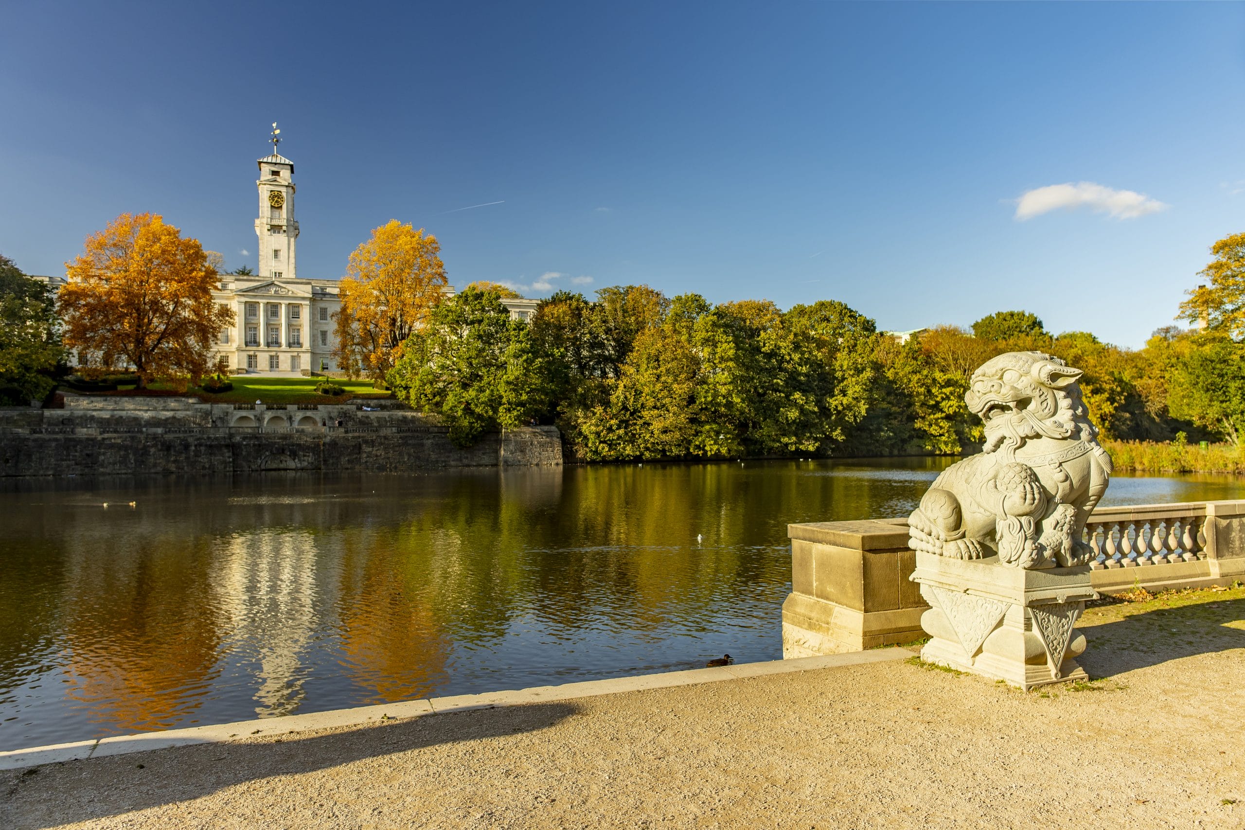 Autumnal trees reflect on a lake with a tall clock tower in the background and a stone lion in the foreground