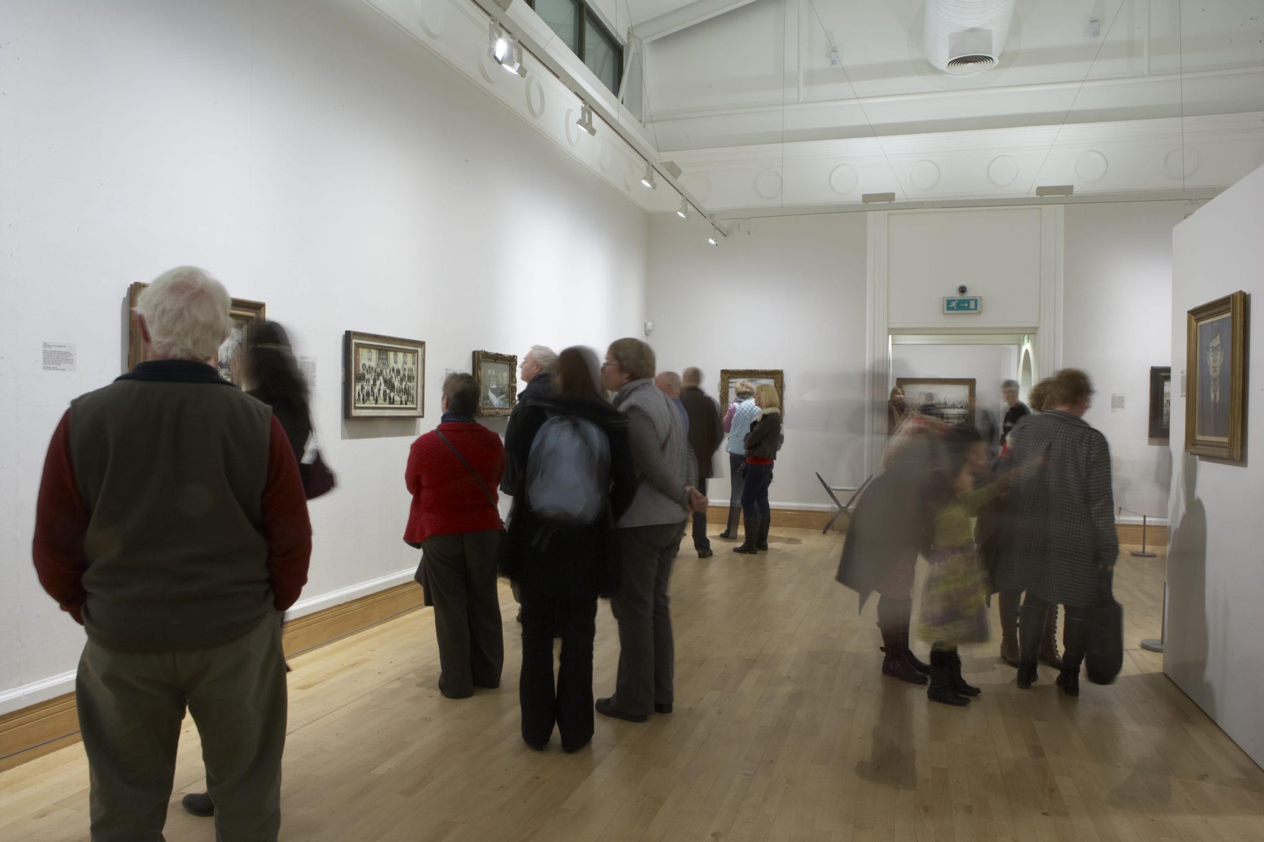 A group of people look at Lowry Paintings in the Djanogly Gallery