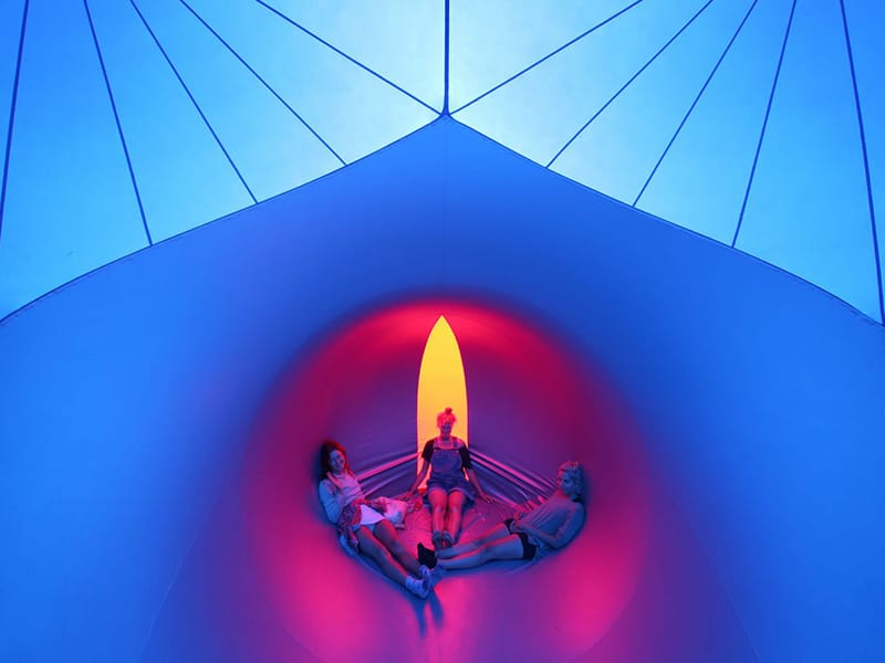 Three teenage girls sitting in small chamber inside the Exxopolis luminarium, backlit by yellow, and surrounded by blue light, with a lighter blue section above with dark blue stripes.