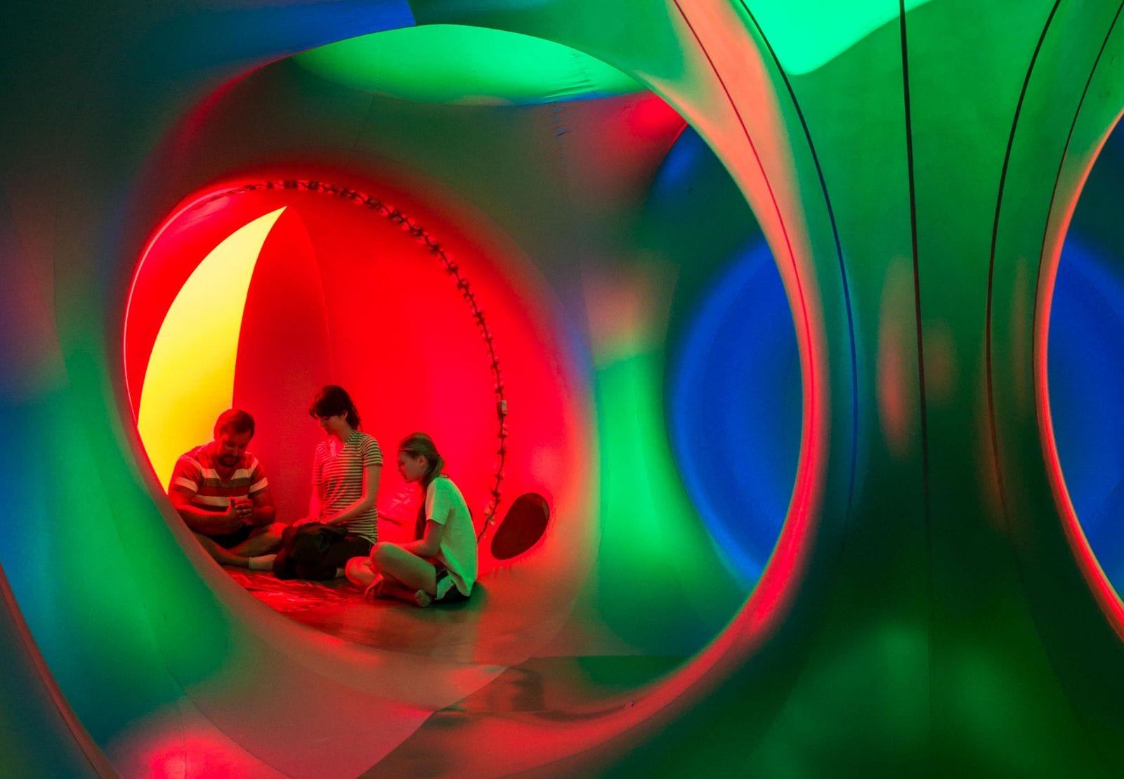 A man, woman and teenage girl sit and talk in a small bright red dome in the luminarium. They are surrounded by towers and walls that glow bright green.