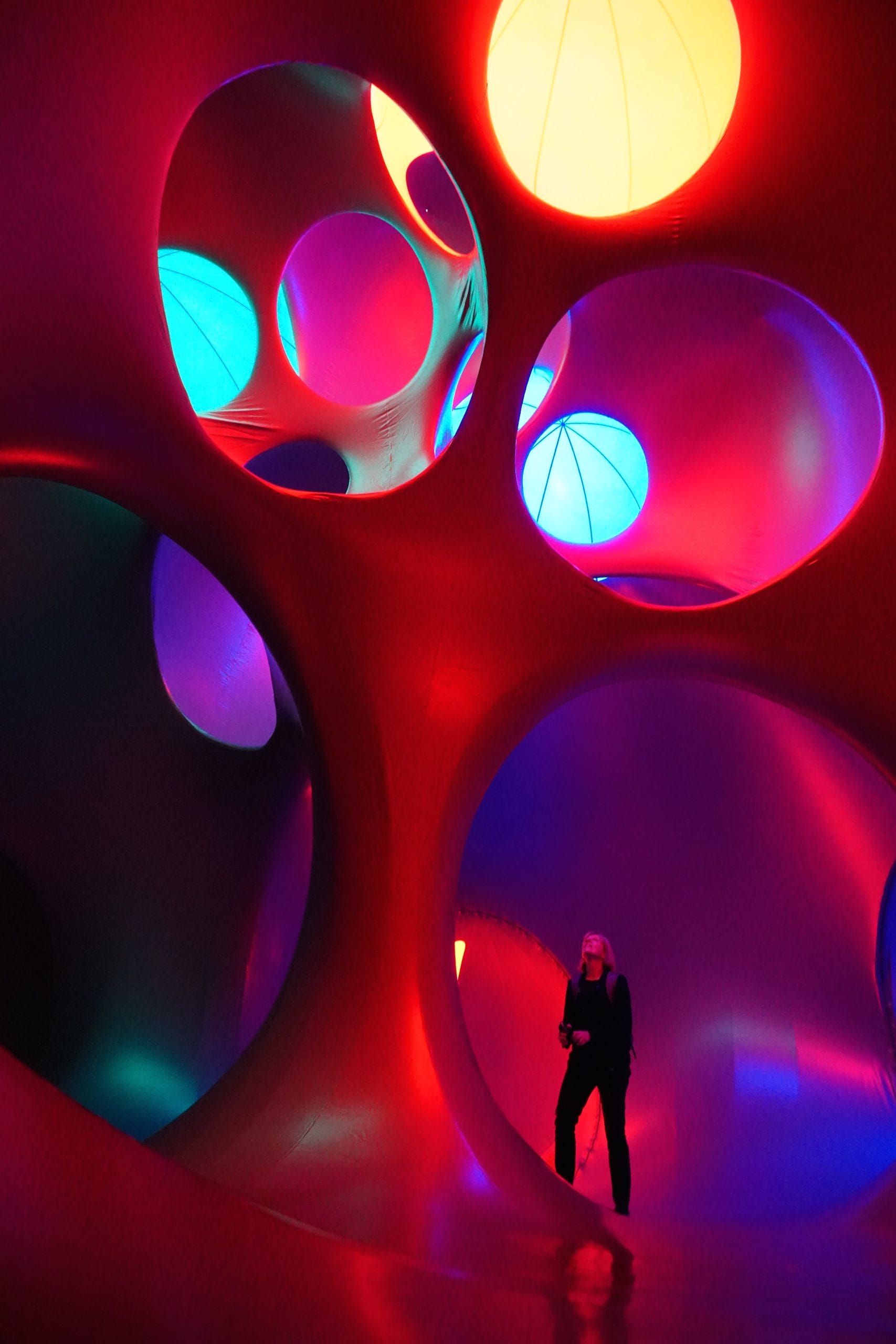 A woman looks up at a luminarium tree, which glows red, blue and yellow and is a series of circles that seems to hang in the roof of a large dome within the luminarium.