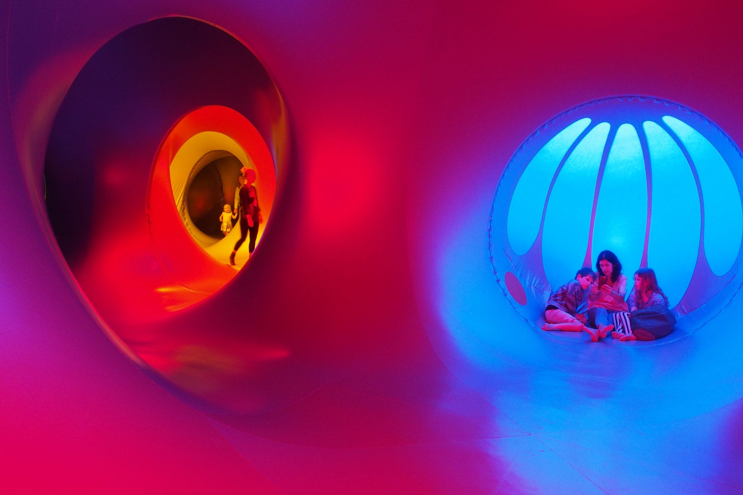 A family sit, cuddle and relax in a small blue dome inside a luminarium. In the background a small child holds her mother's hand whilst exploring in a bright yellow section.