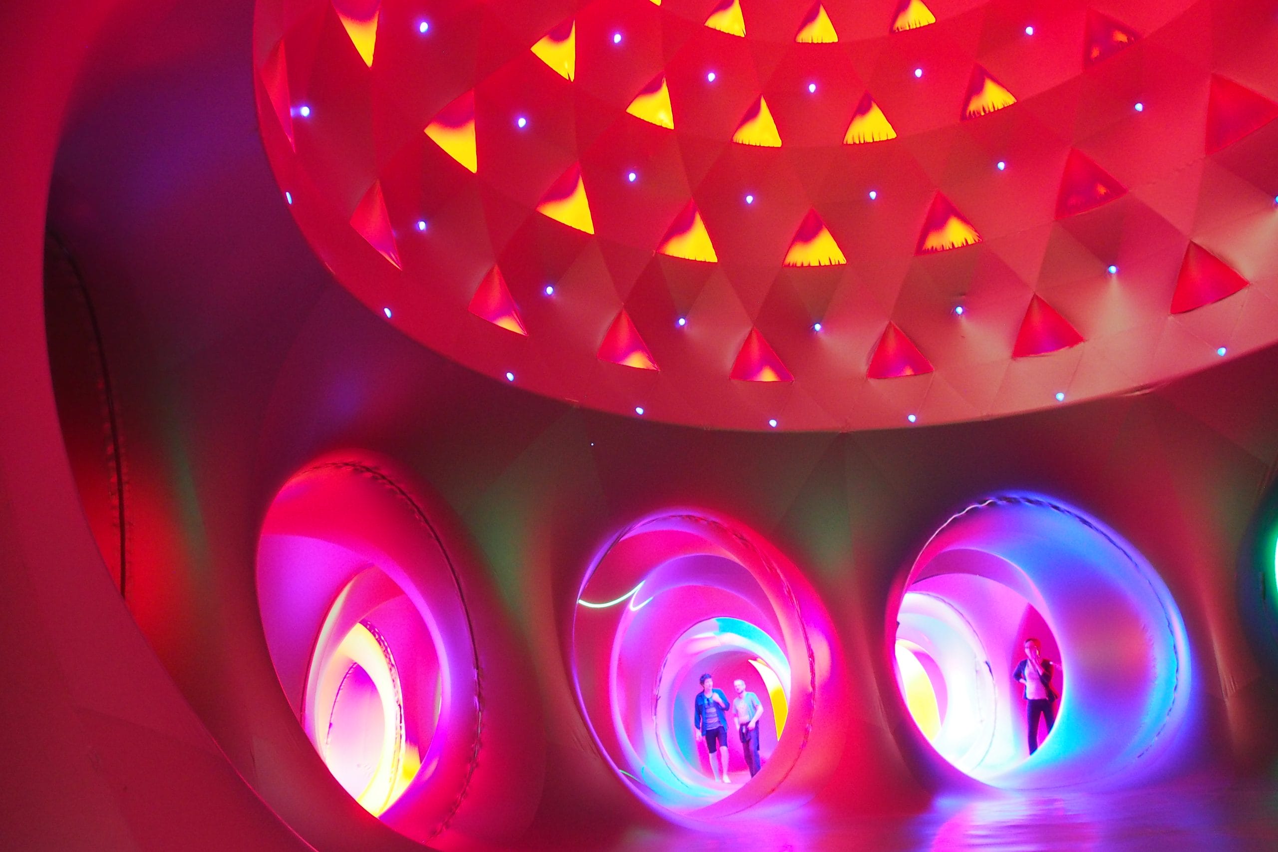 A larger dome within the Katena luminarium with geometric shapes in the domed roof. Predominently red, tunnels branching off from the dome are purple and blue. People explore the tunnels in the background.