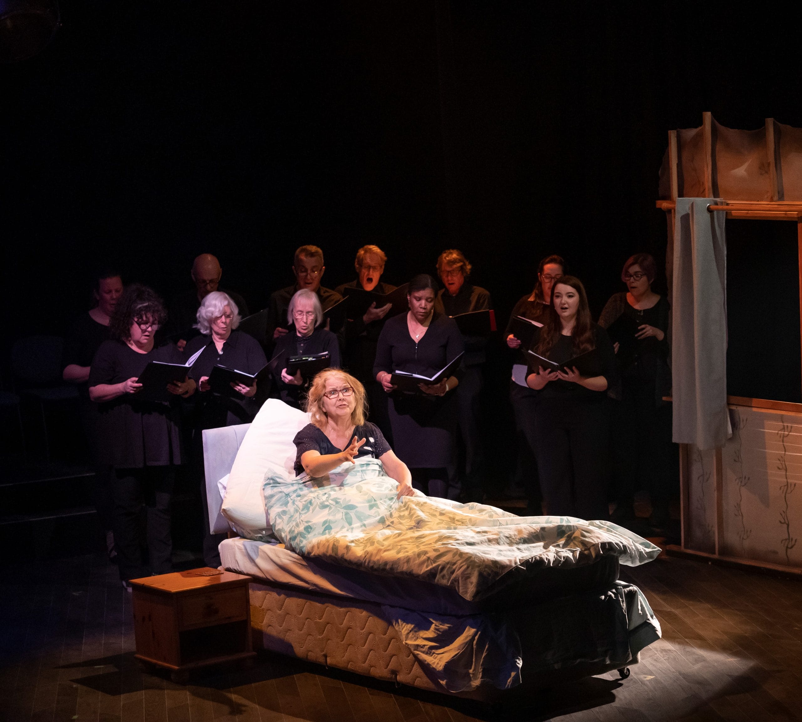 A woman in bed reaches out as a choir sings around her