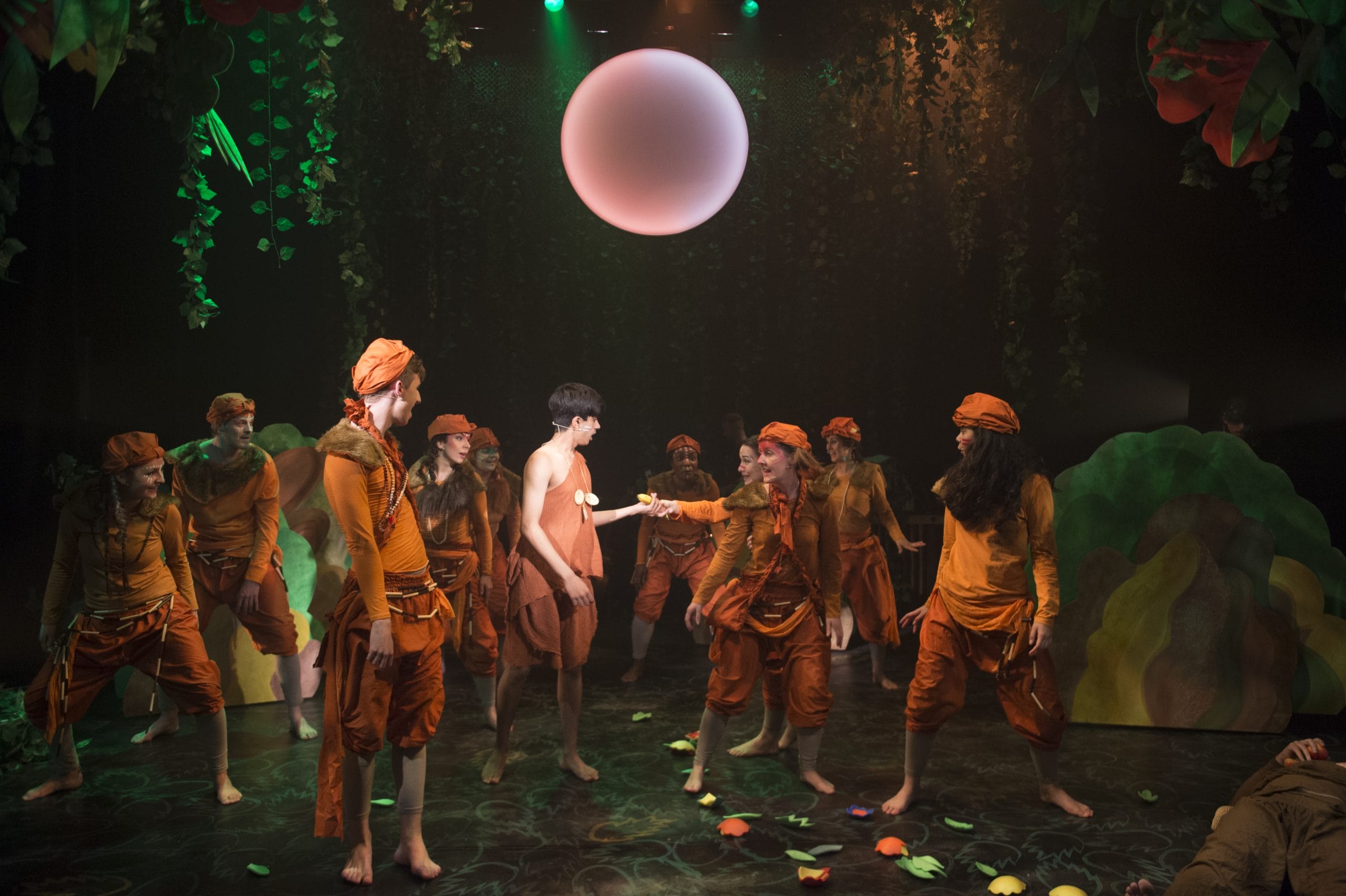 Eleven actors on stage in orange costumes and in a jungle scene, with the moon overhead.