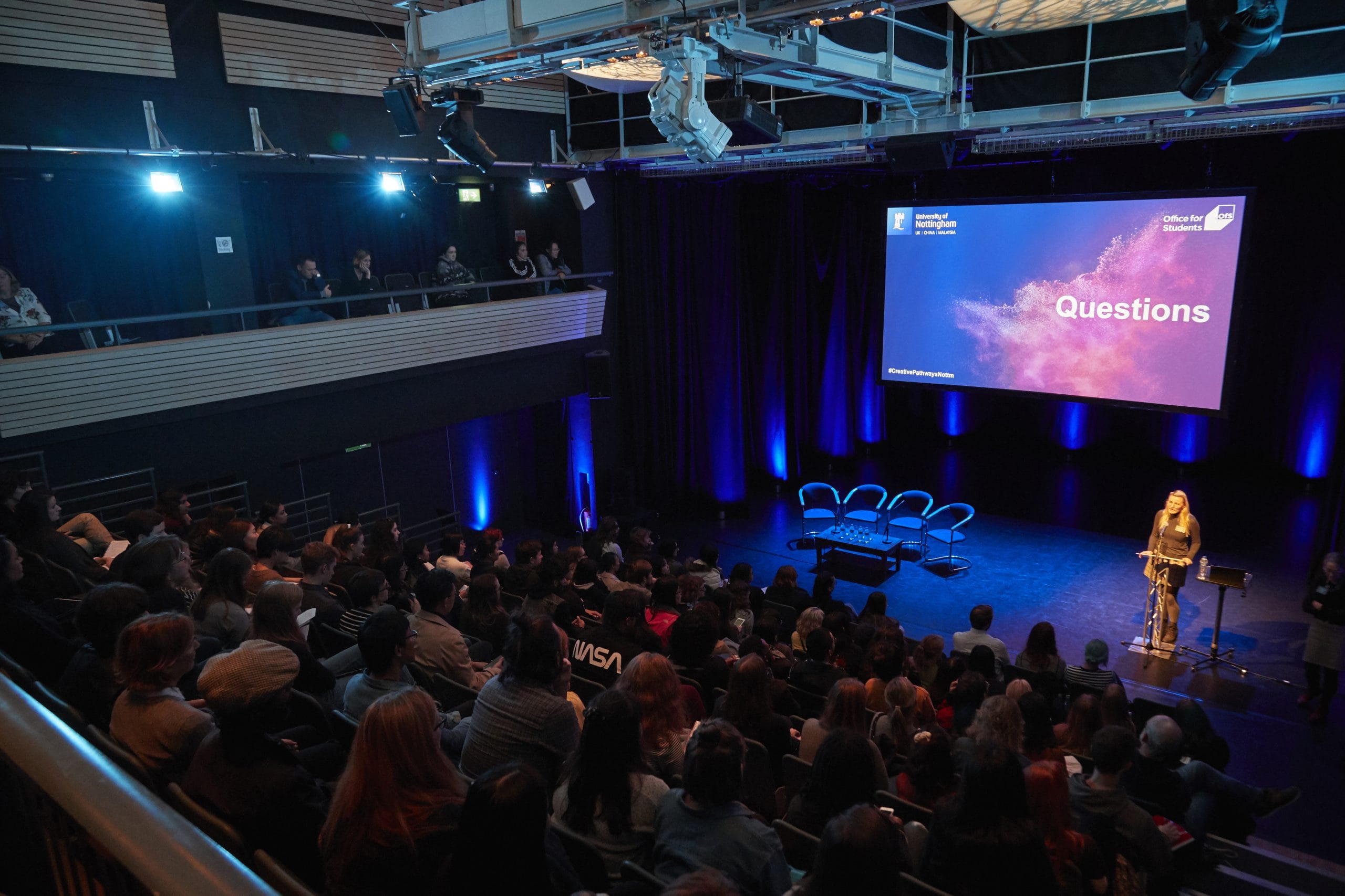 Lakeside's theatre with a full audience, a speaker at a lecturn and four empty chairs on stage.