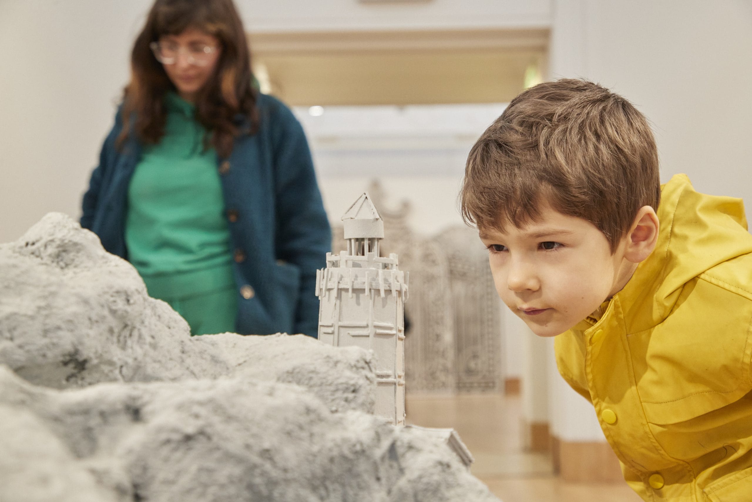 A small boy in a yellow coat admires a rocky landscape sculpture in a gallery, with a lady in a green jumper in the background.