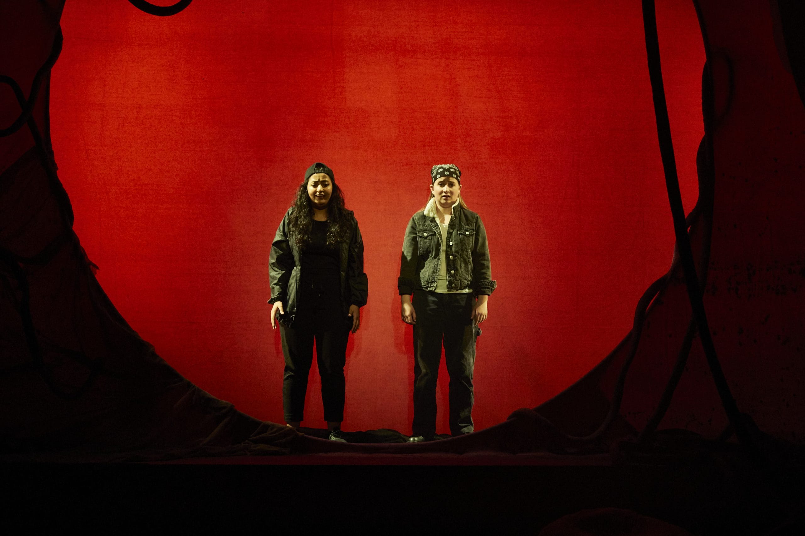 two actors stand on stage looking distressed at the audience with a red circle background