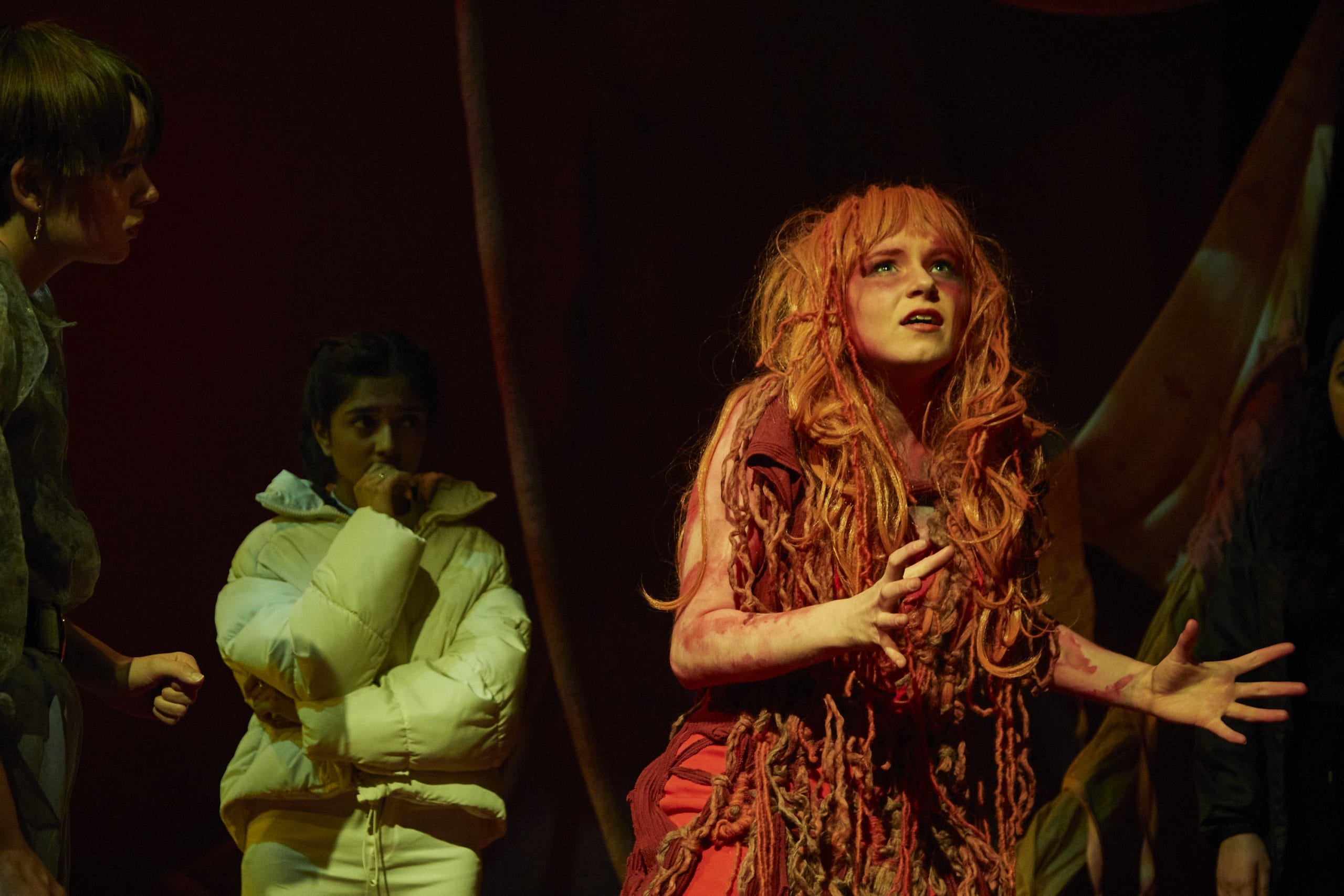 From a production of DNA, a dishevelled looking actor in a red wig and red ripped clothes stares out at the audience whilst other actors look nervous behind her