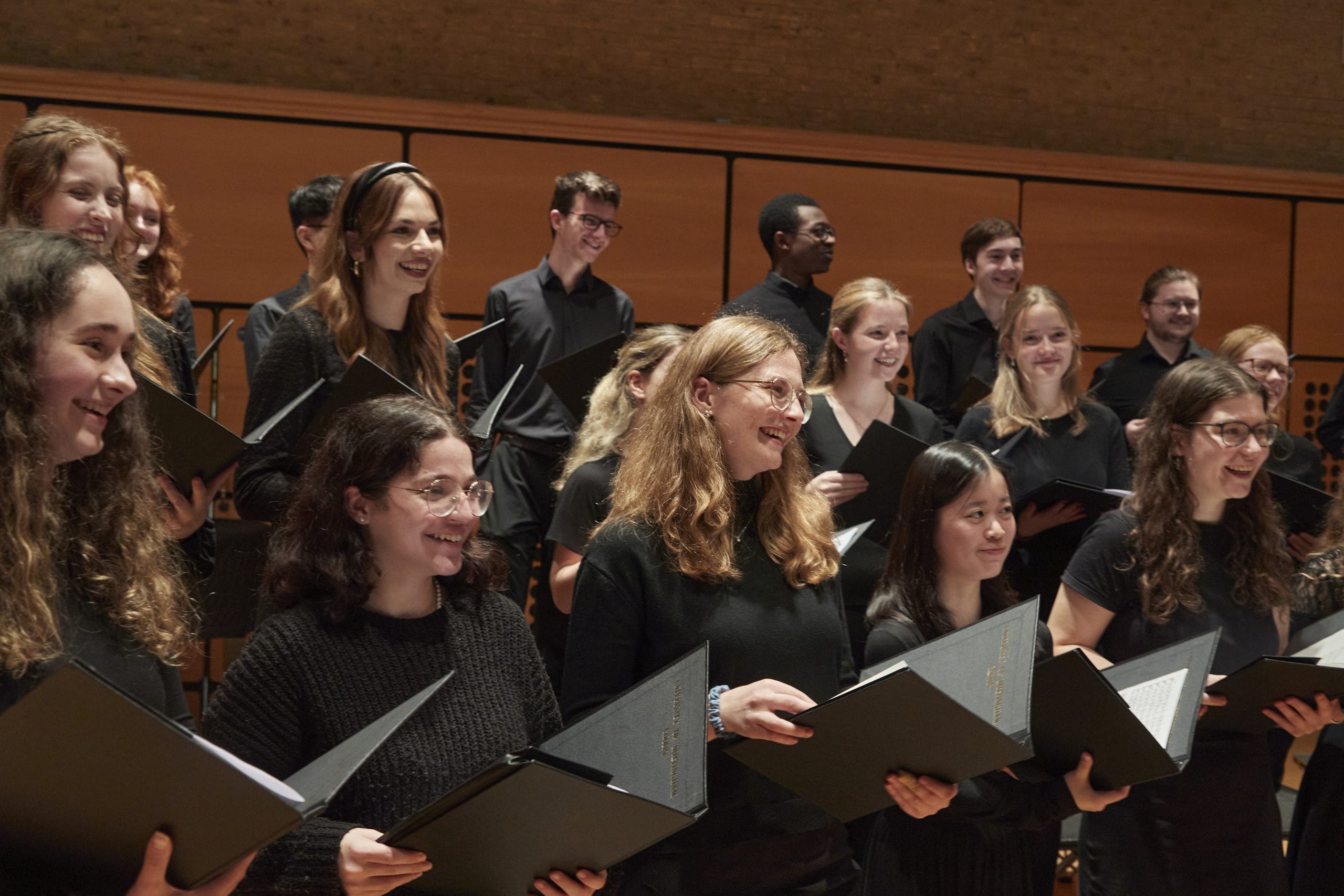 A group of student choir members smile at their director holding song books