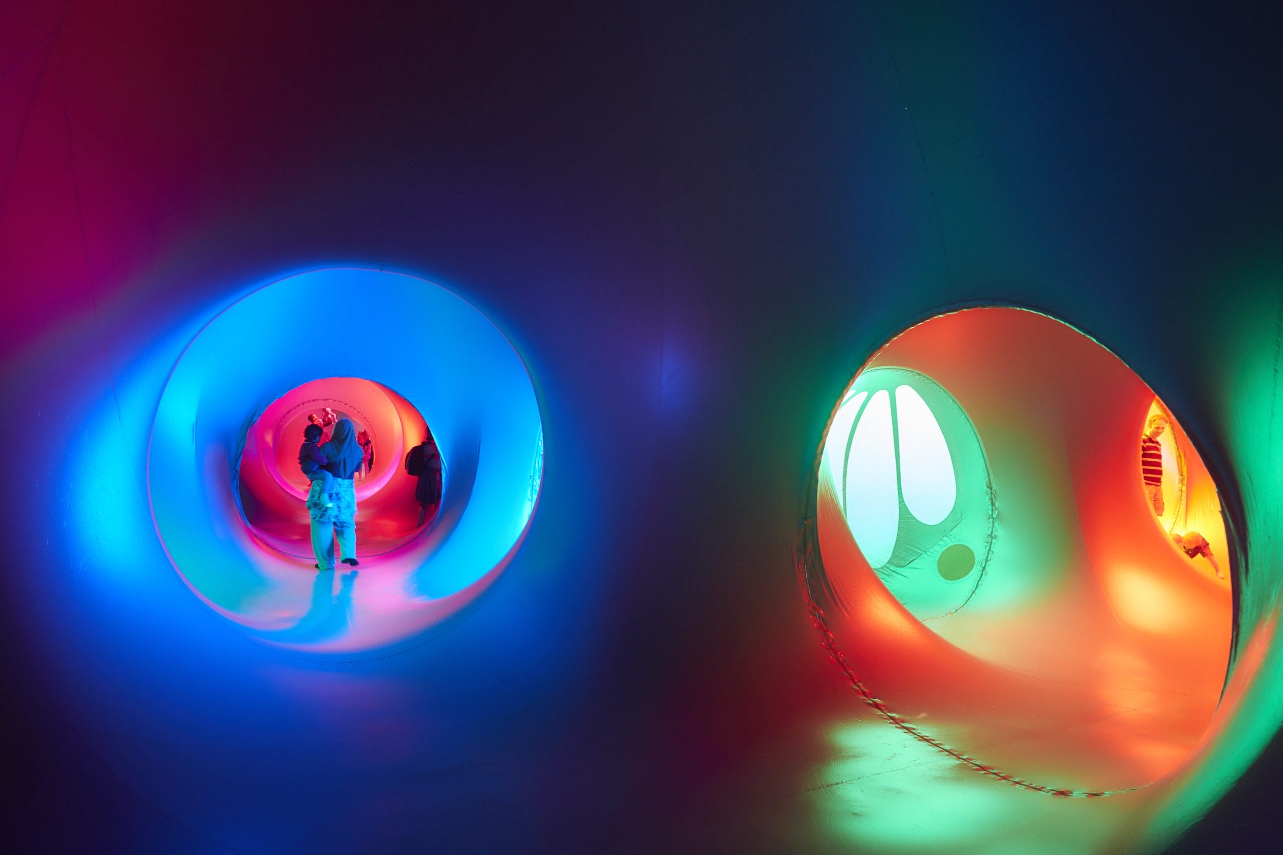 A lady with her back to the camera carries a small child through a tunnel in the luminarium. The tunnel glows blue, with reds and pinks further ahead. To the right, greens, reds and golds can be seen in another section of the luminarium, with a man and child seen emerging round the corner.