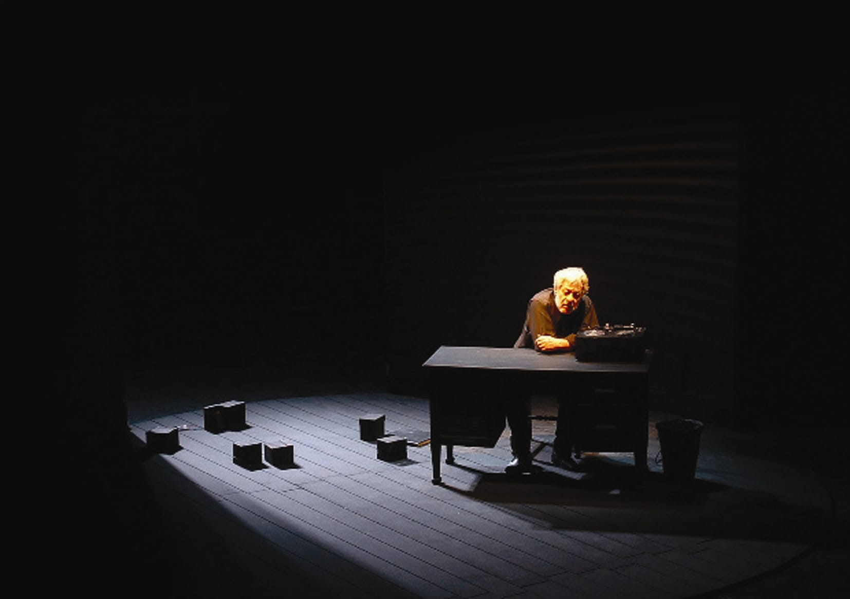 A man sits on stage in the dark with a table and lots of tapes scattered around him