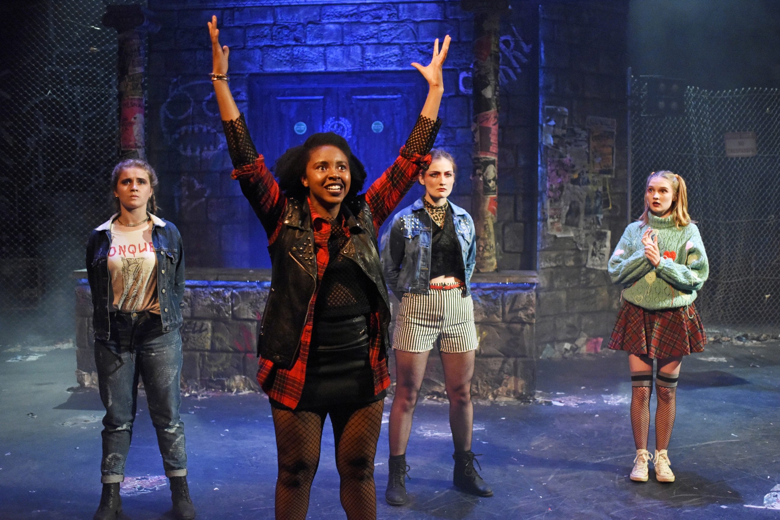 three actors stand like soldiers whilst a fourth raises her arms in glee