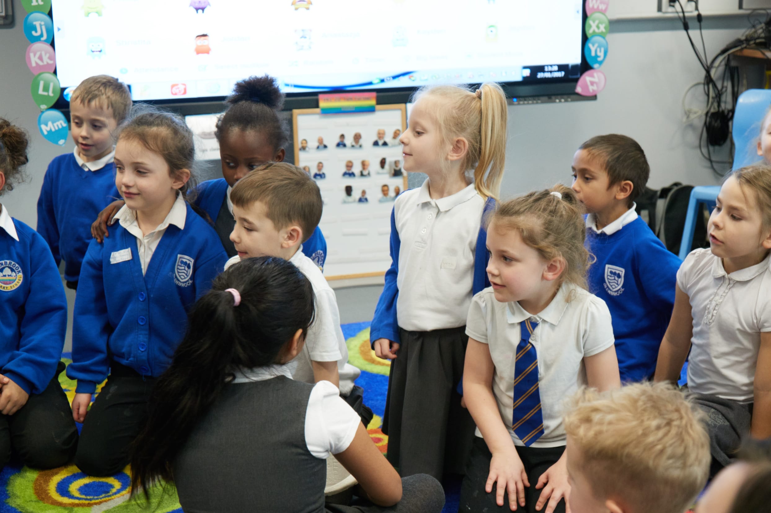 A dozen primary school children in blue, white and grey uniforms smiling and looking inquisitively at something out of shot of the image.