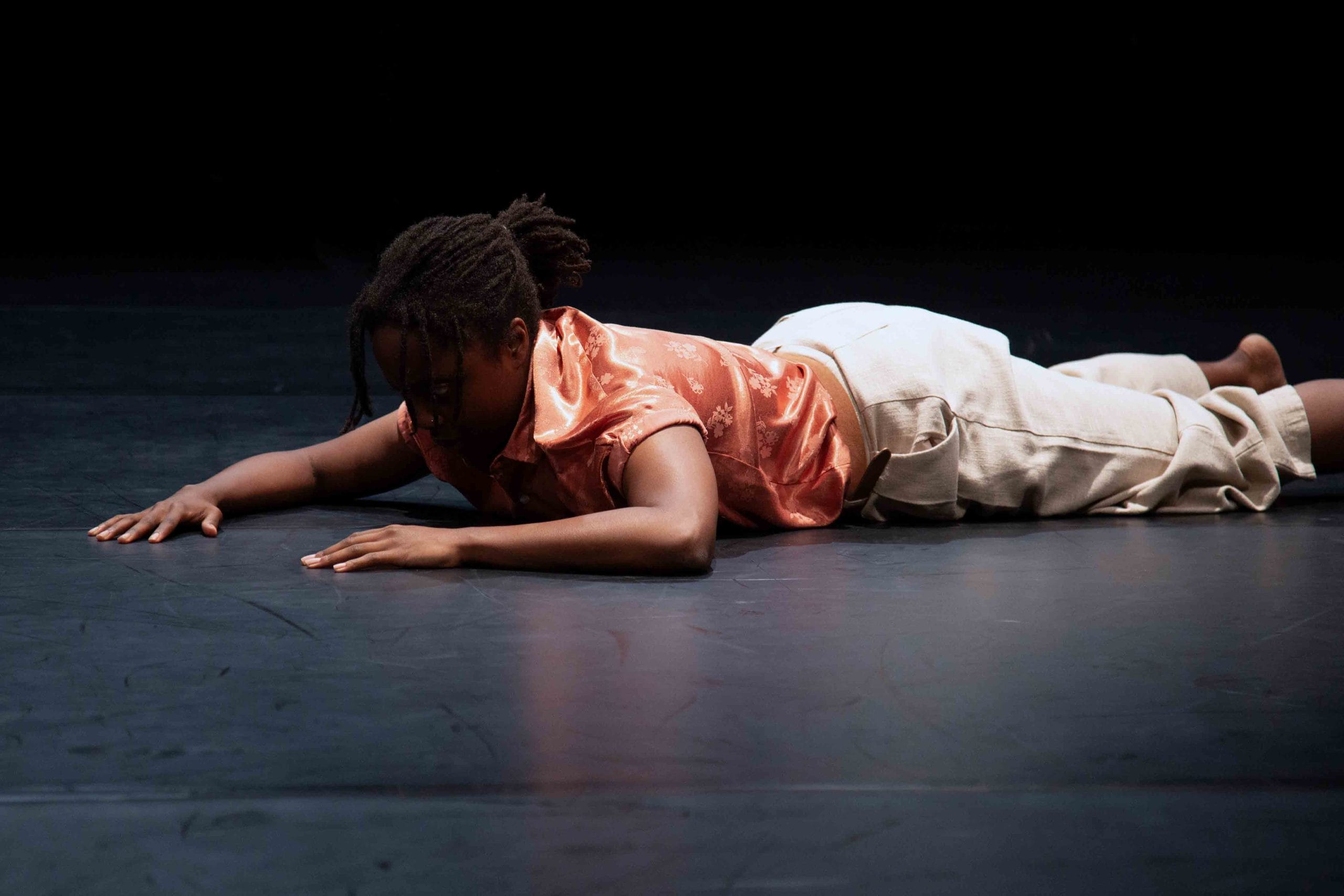 A dancer in a pink top and white trousers lies on stage