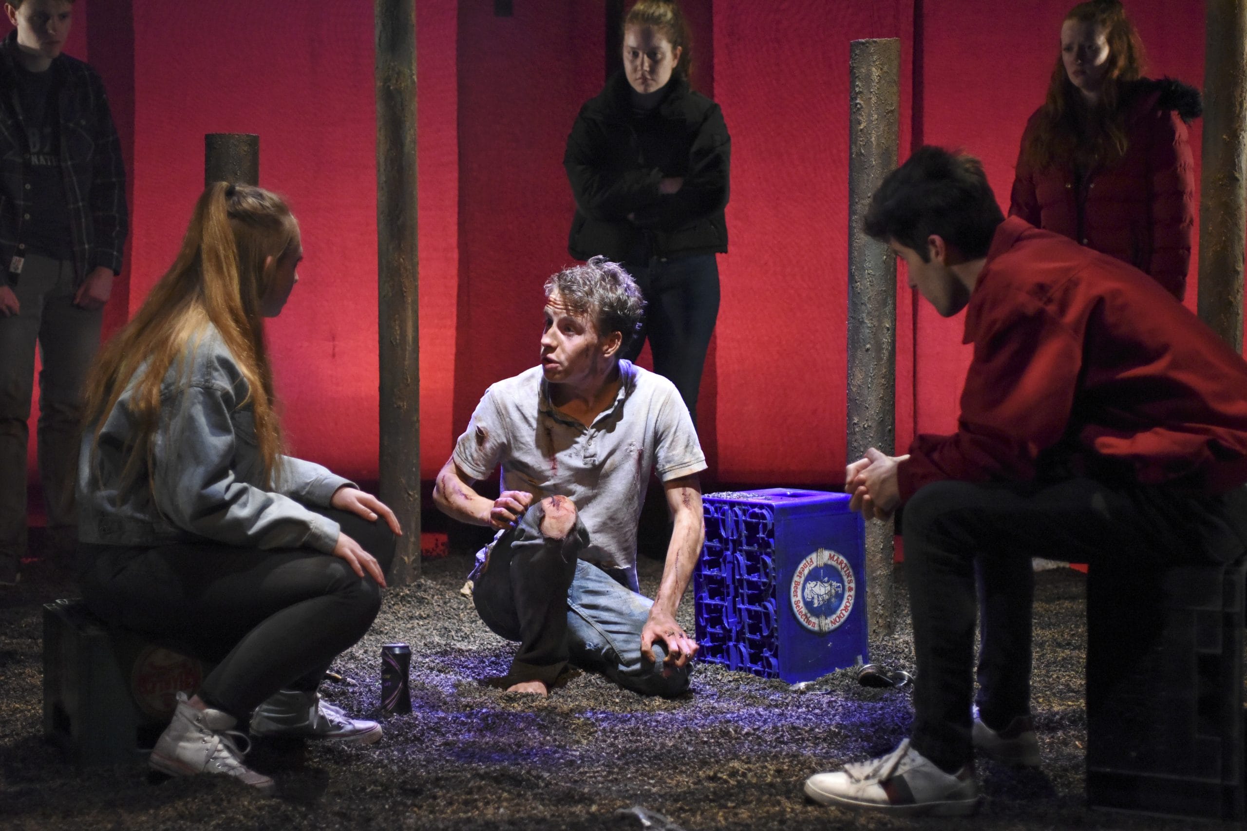 a group of actors look at a dishevelled man in a circle, there isa red background and pillar around them