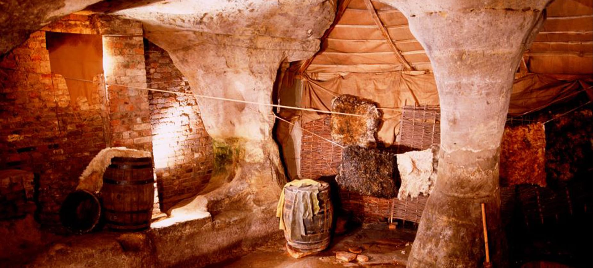 The inside of a cave with animal skins hung up and barrels of dyed fabrics
