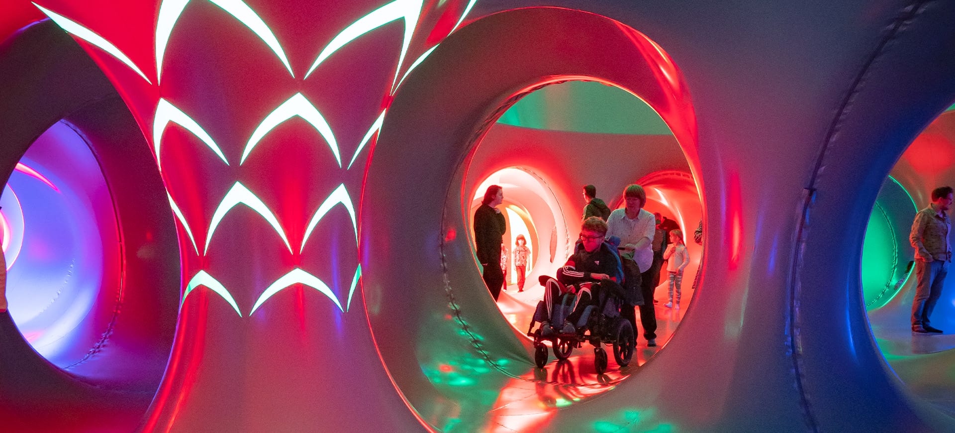 Mum pushing boy in wheelchair through luminarium, which is glowing shades of pink, blue and green.