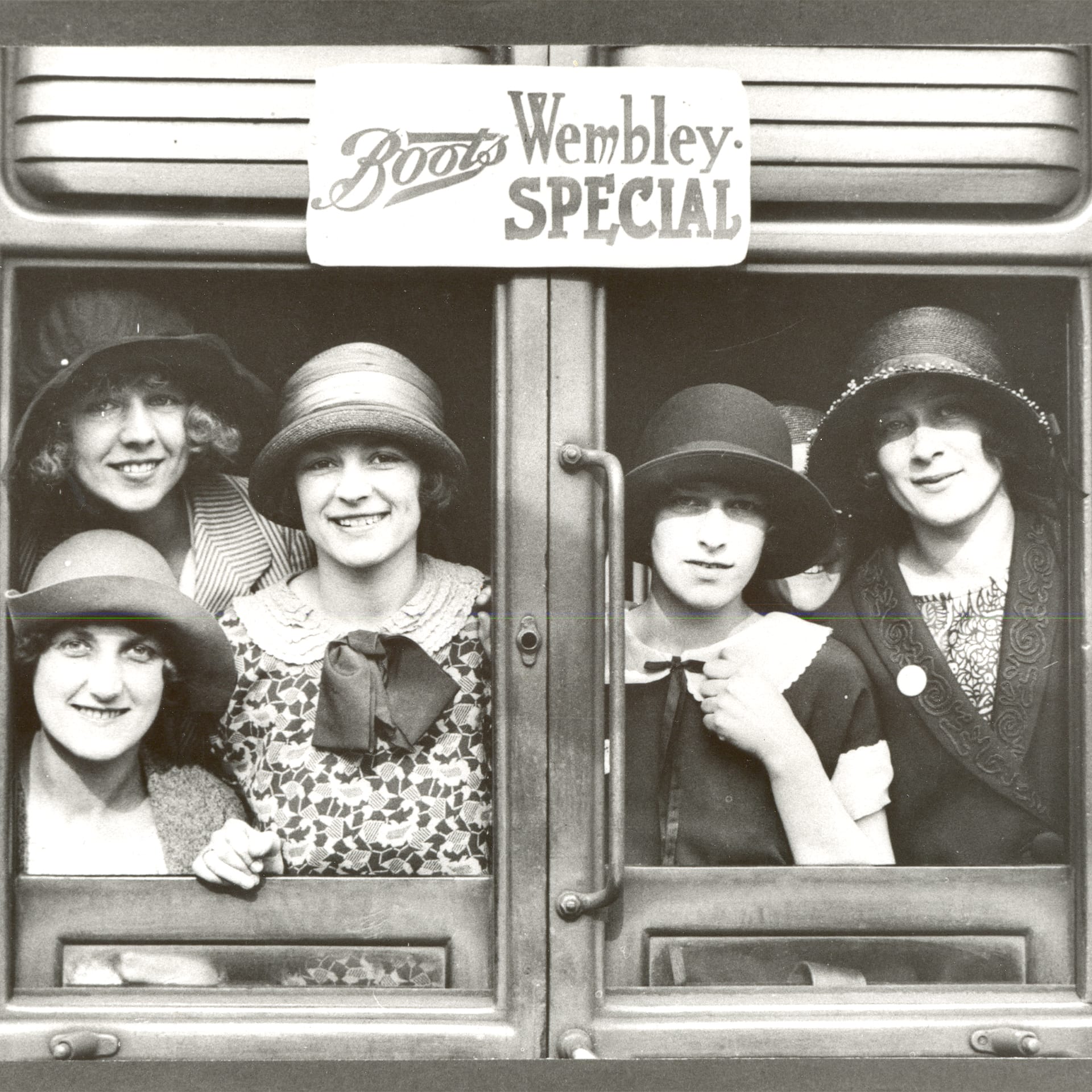 A black and white photo of a group of women in hats underneath a Boots sign that reads Wembley Special