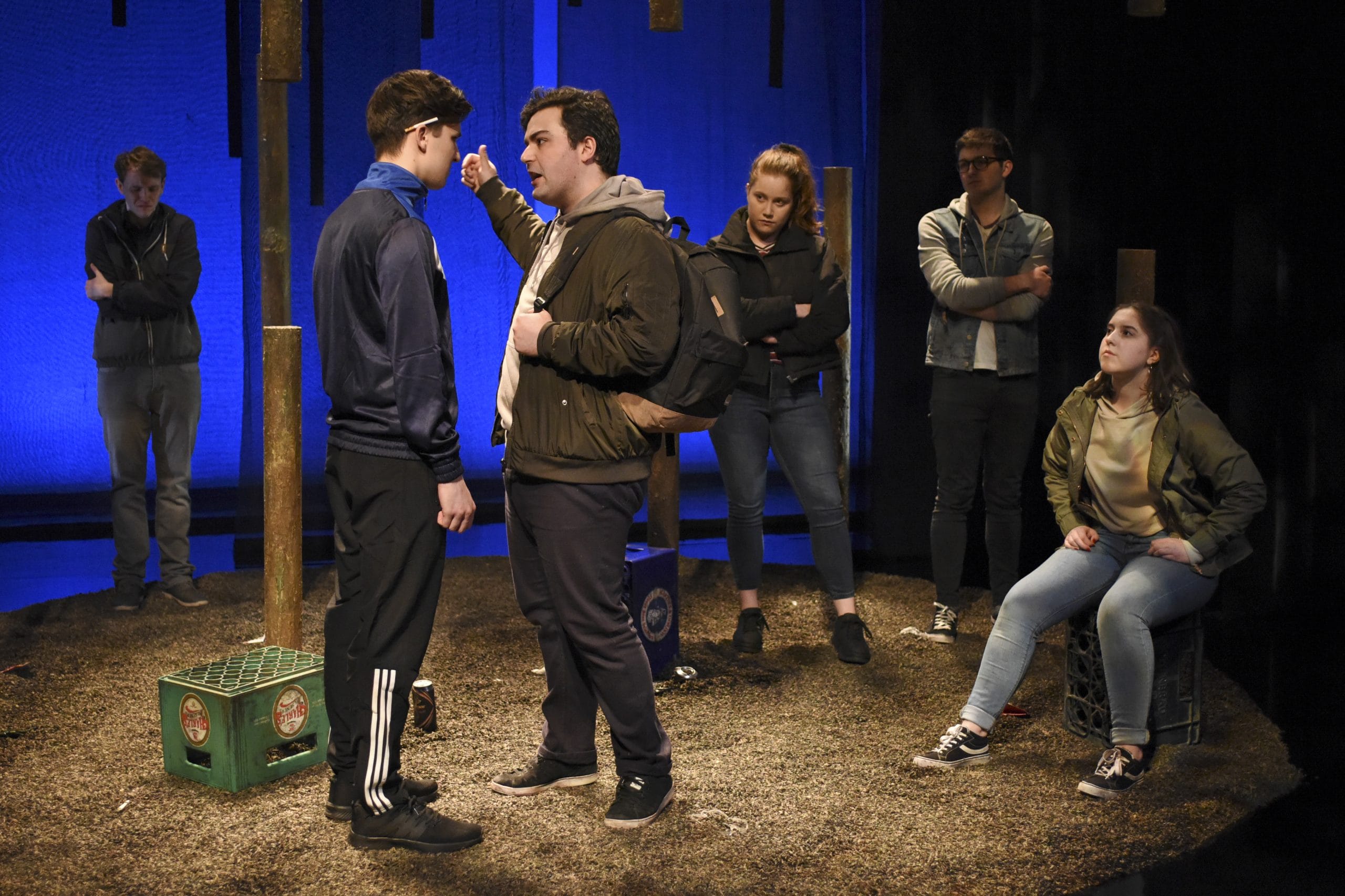 two actors fight whilst four other actors watch them with arms crossed