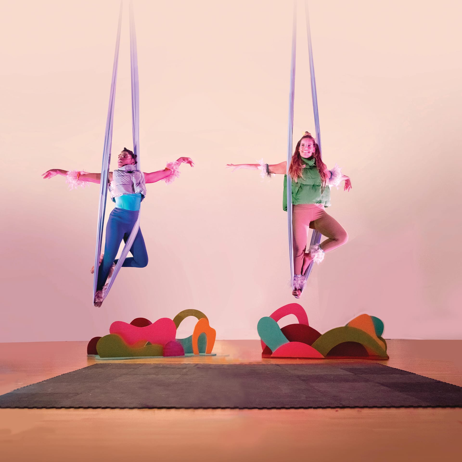 Two women with feathers on their arms fly high above a stage with silks