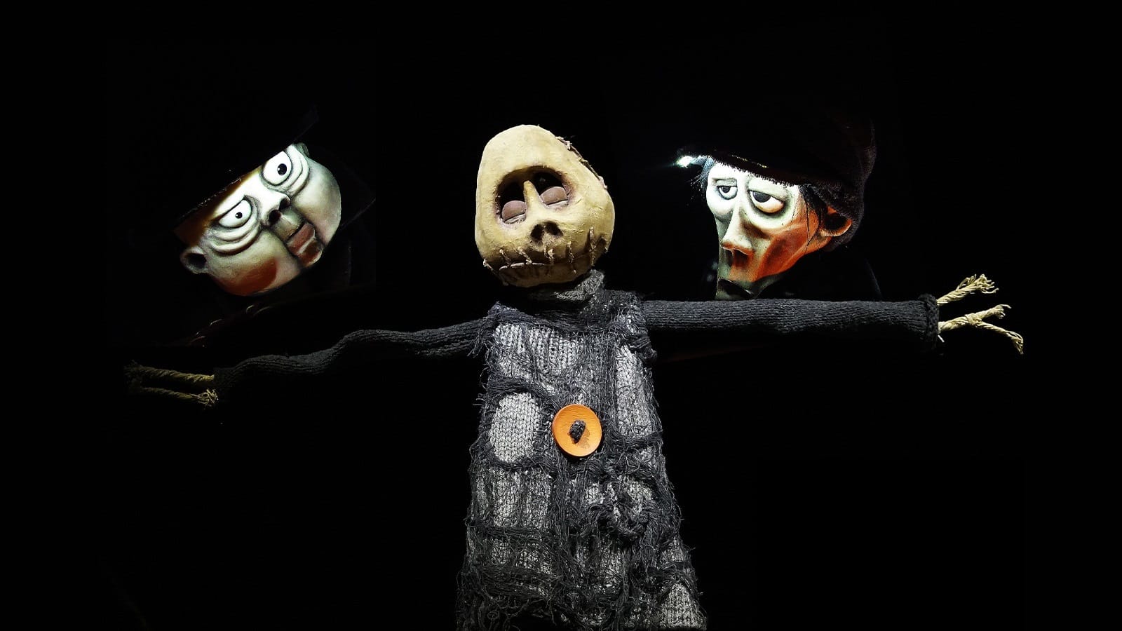 A scarecrow puppet looking down as two men puppets glare at it