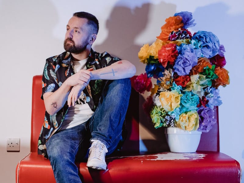 A man with a colourful shirt and a brown beard sits on a red chair with some colourful flowers next to him in a vase
