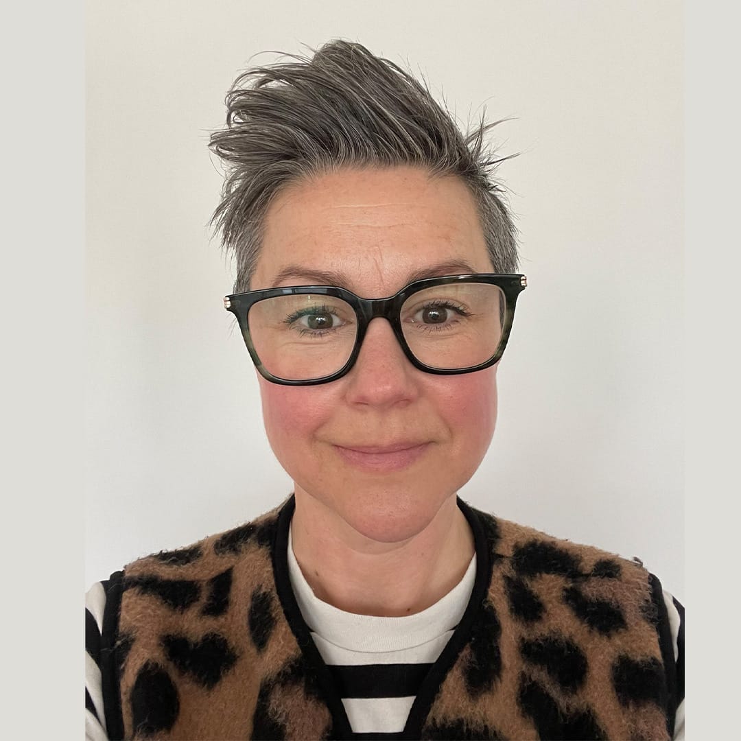 A woman with short hair and glasses wearing an animal print gilet.