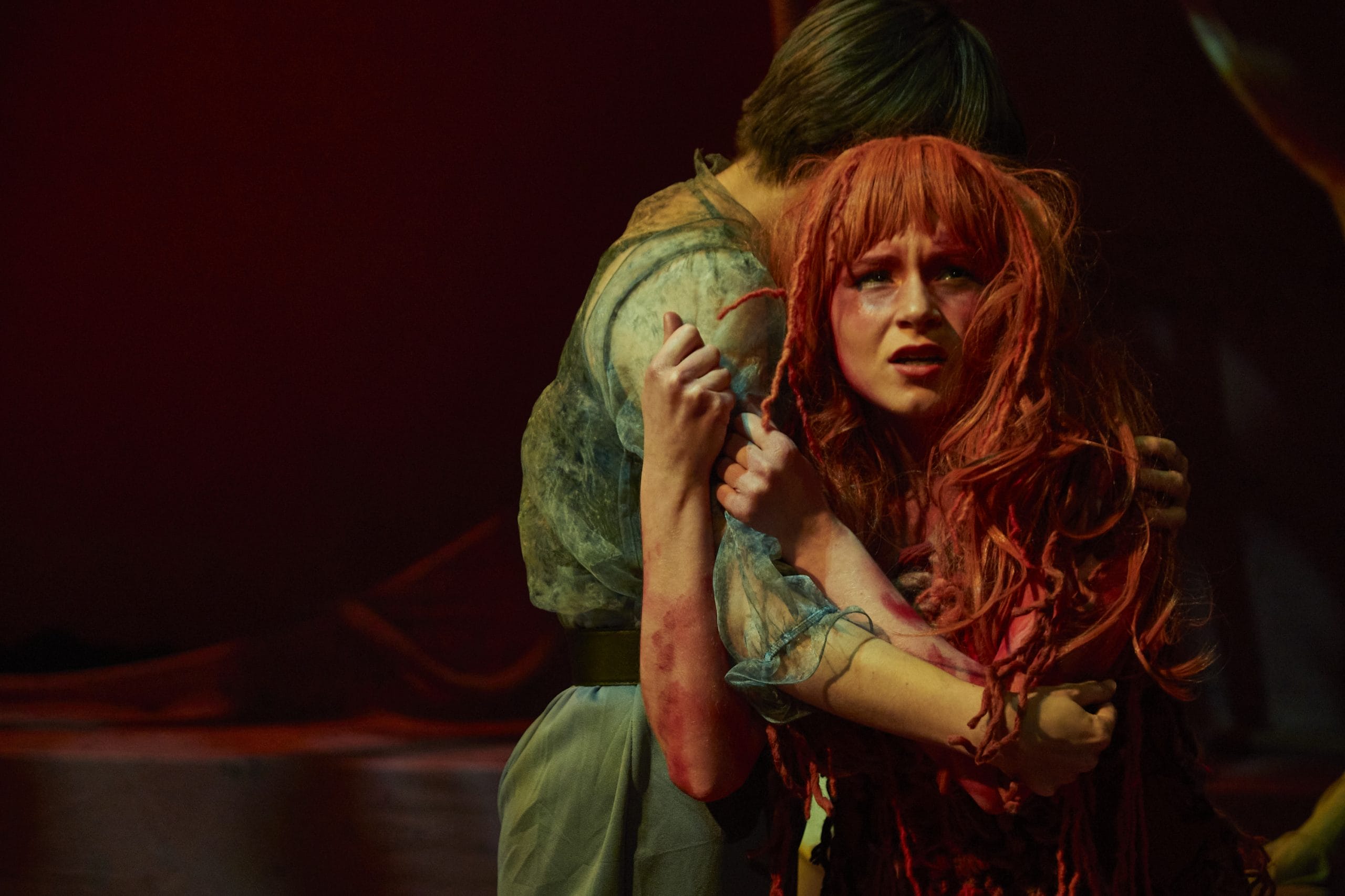 A student actor on stage with red hair being led off by another student