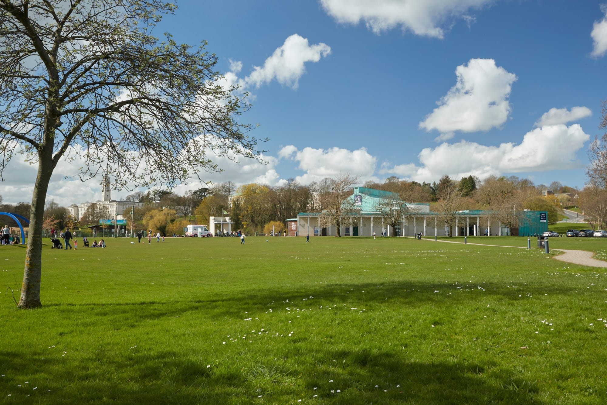 Grass interspersed with daisies, a winding path and a small tree just coming into lead dominate the foreground. Children playing in Highfields Park, an ice cream van and Lakeside Arts' DH Lawrence Pavilion are in the background. A bright blue sky with white fluffy clouds is overhead.