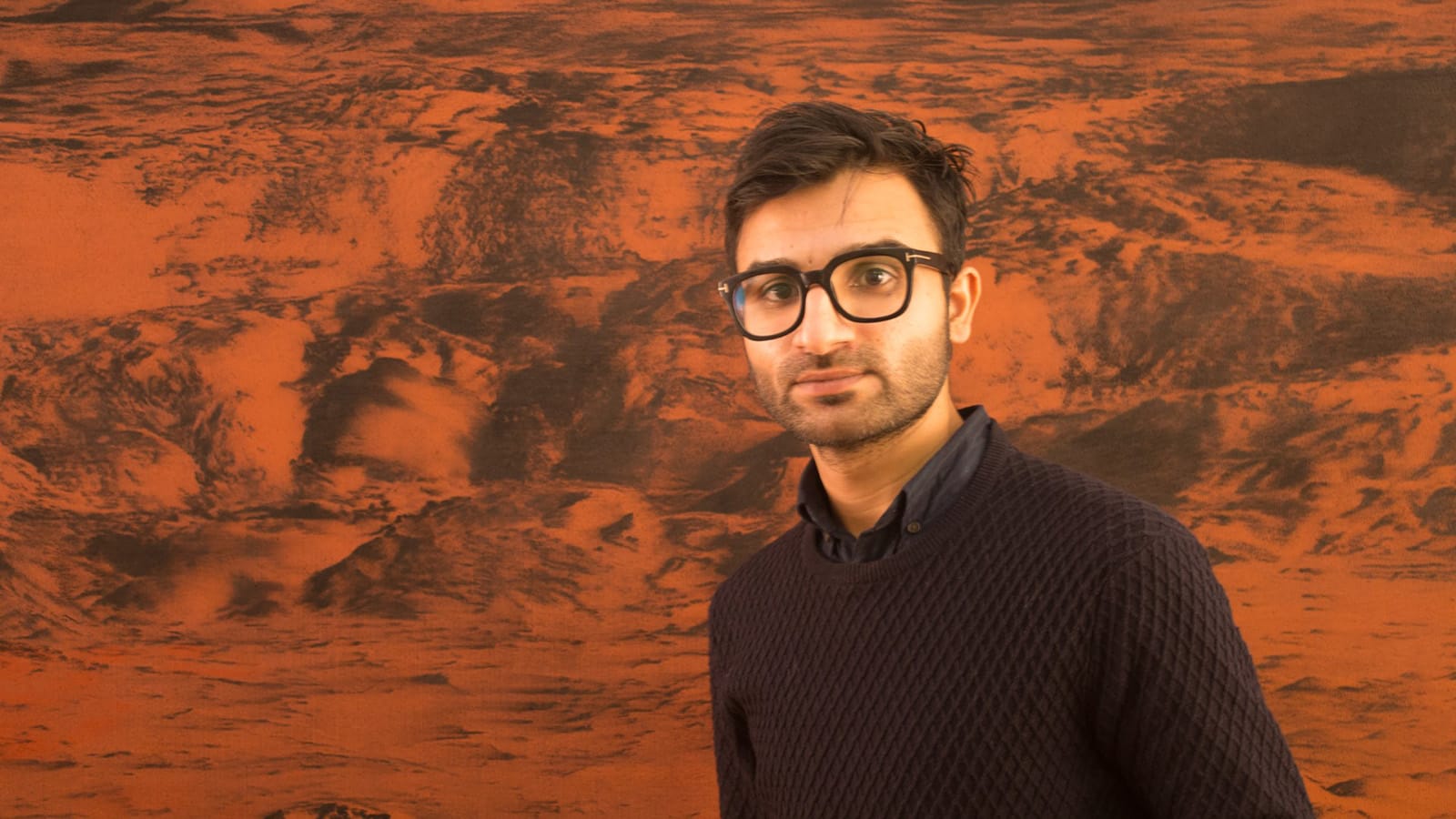 A man wearing black glasses with dark brown hair, standing in front of a painting