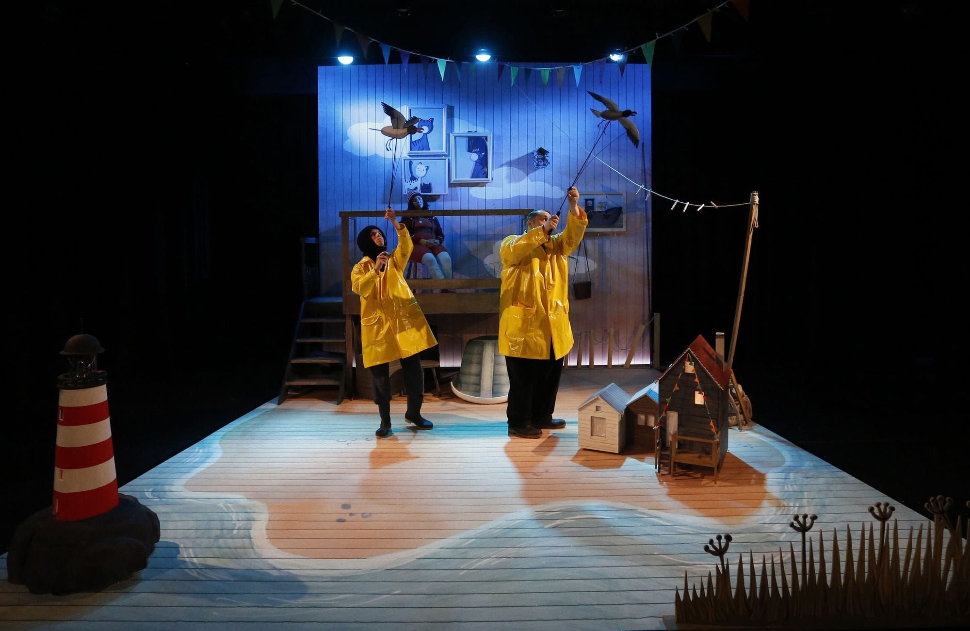 Two actors in yellow raincoats hold up seagull puppets