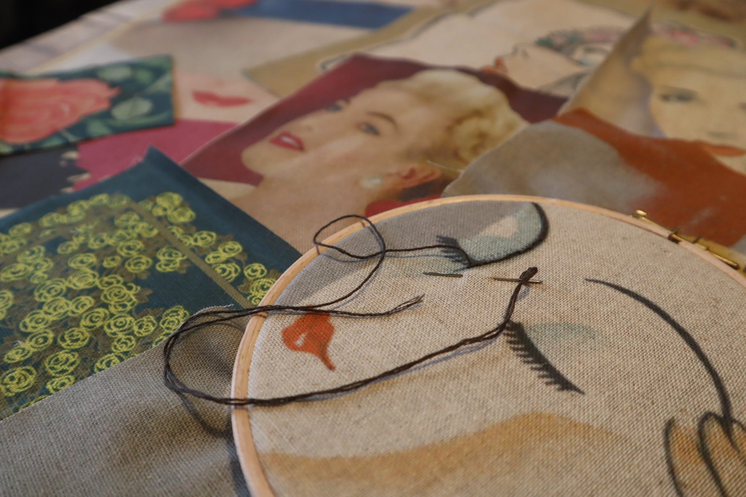 An embroidery hoop, featuring a ladies face, sits on a table surrounded by fabric and pictures.