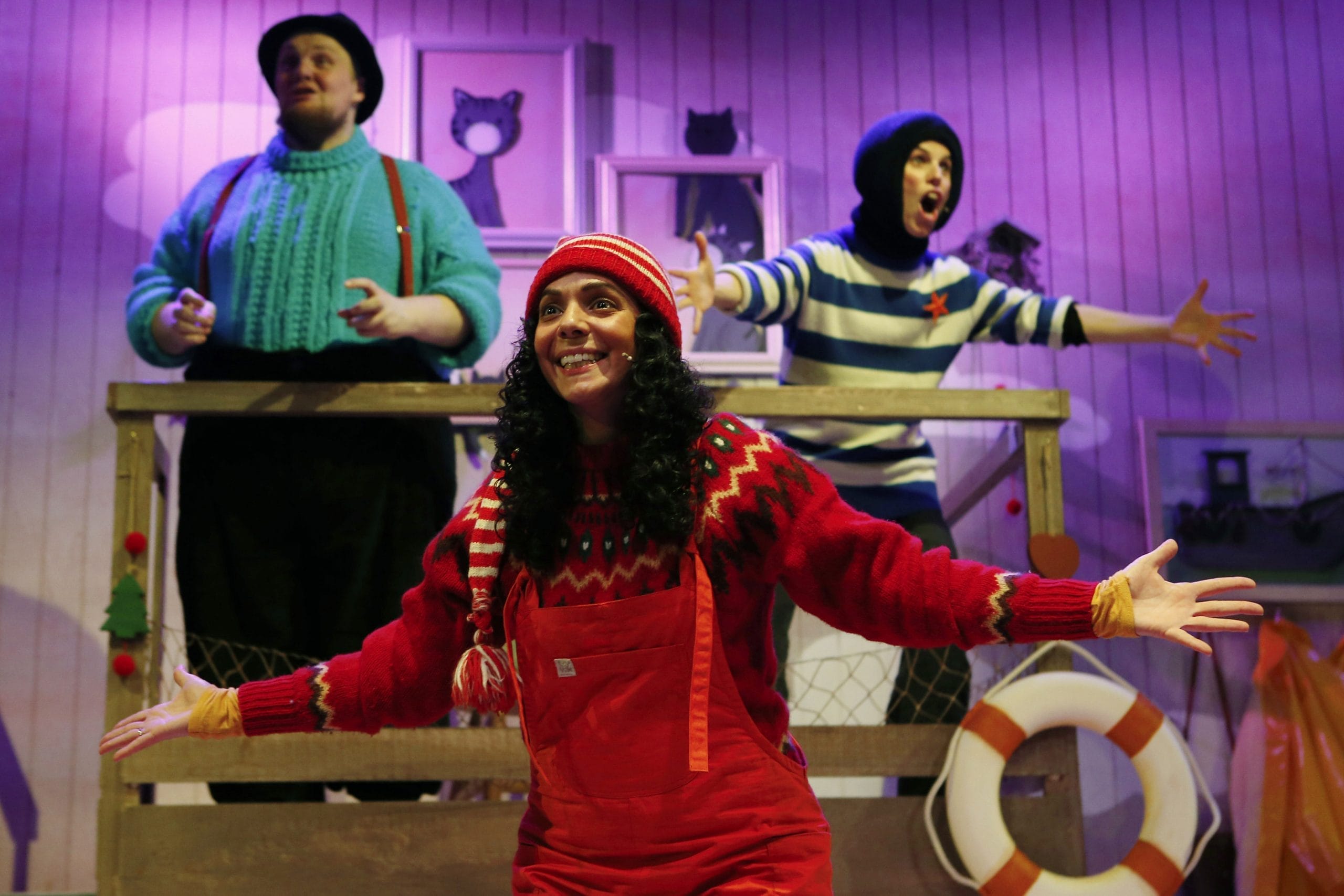 A lady wear red dungarees and a red beanie hat, with long dark hair stands at the front of the stage with two men stood at the back for the stage, one wearing a green shirt and braces, the other in a striped blue and white top.