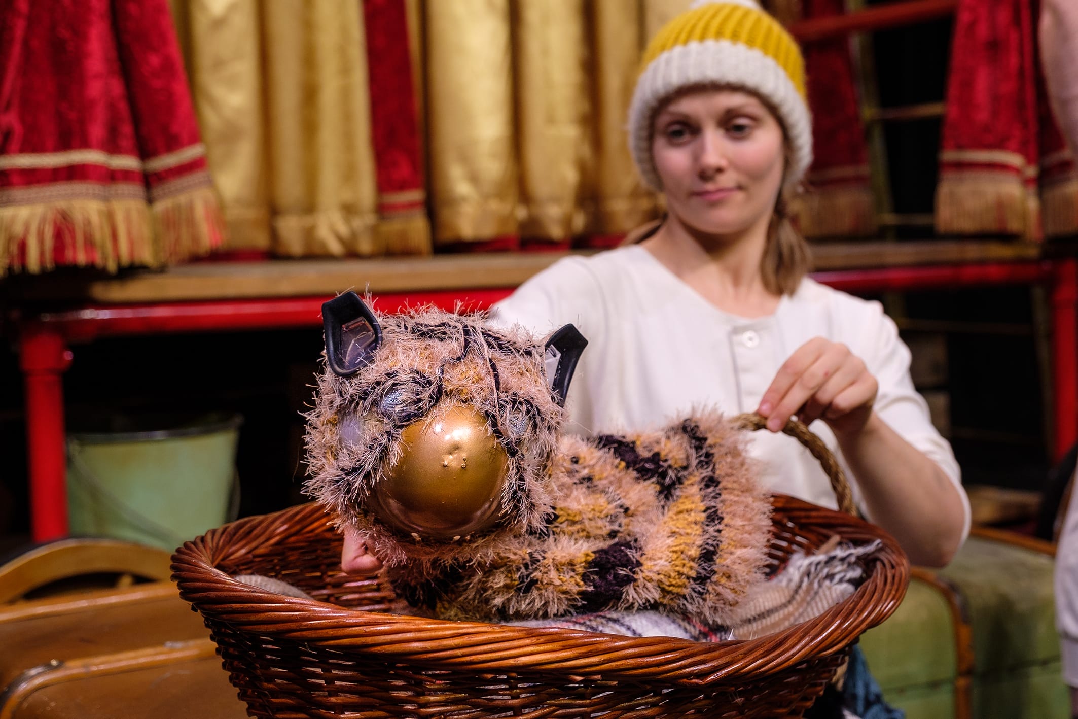 A tiger puppet sits in a wicker basket, with a lady behind in a white top and yellow and grey bobble hat.