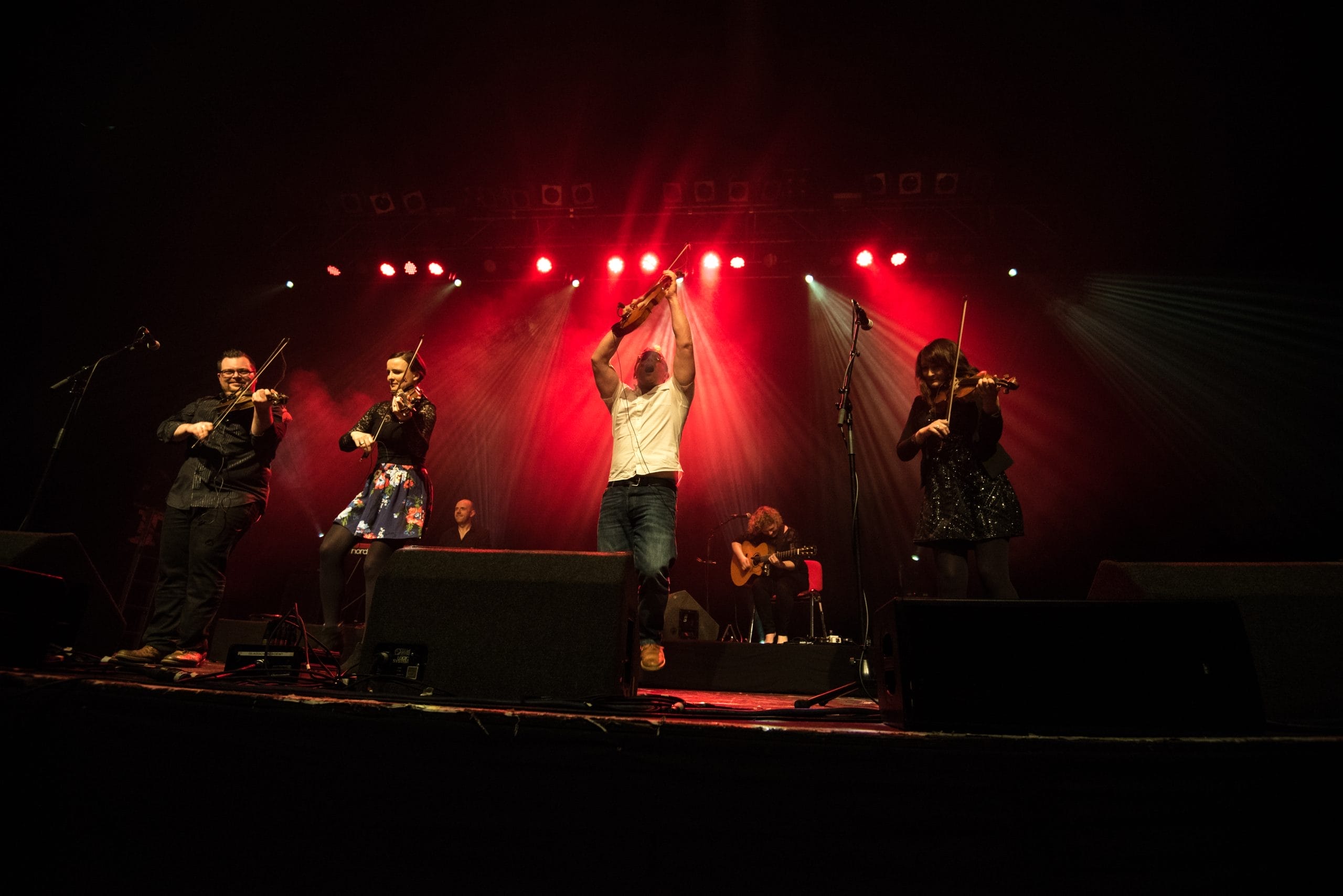 six musicians performing on stage with red lights around them with fiddlers in the foreground