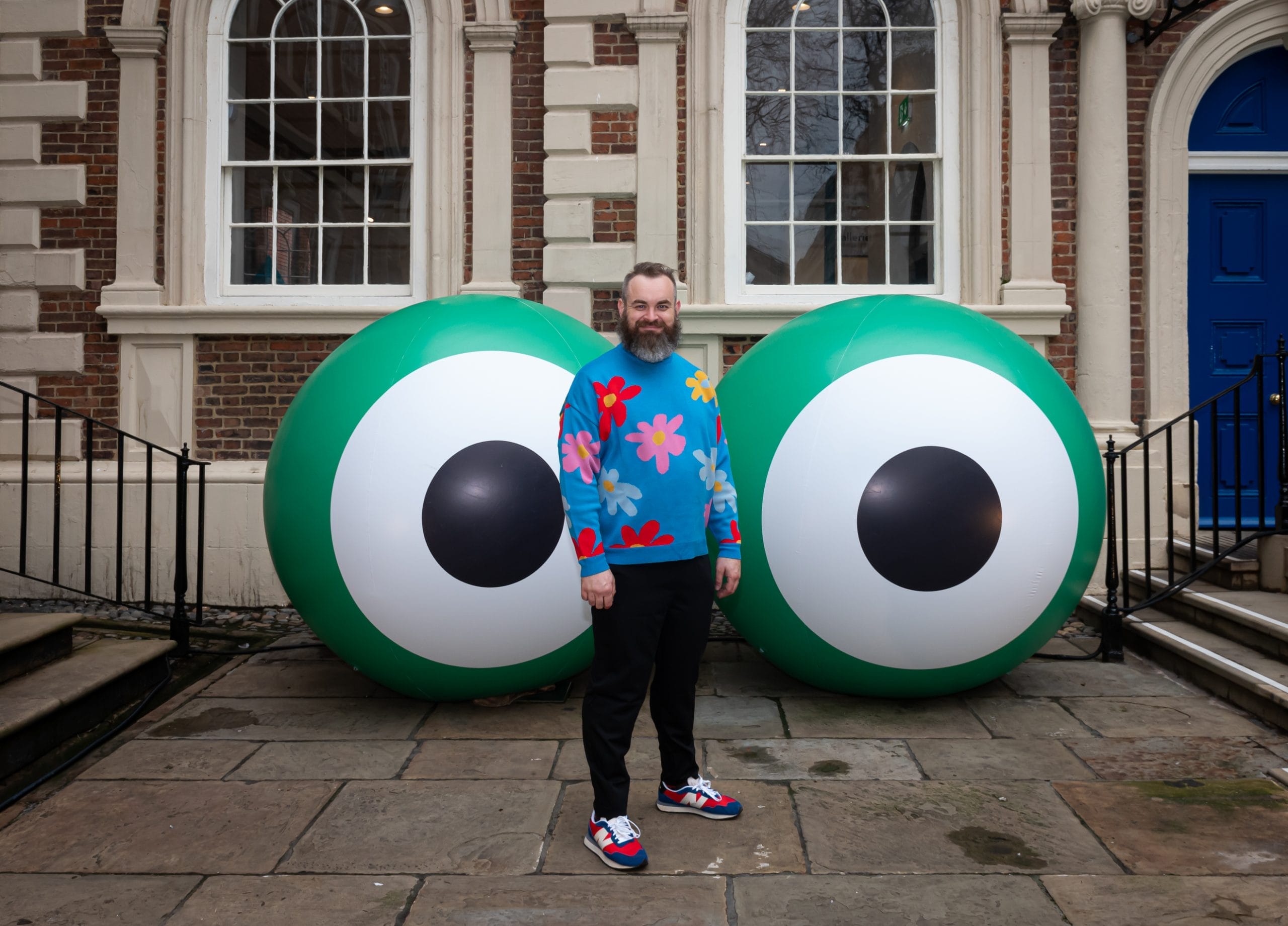 Man in blue jumper with flowers and black jeans standing in front of red brick building with giant green white and black eye inflatables