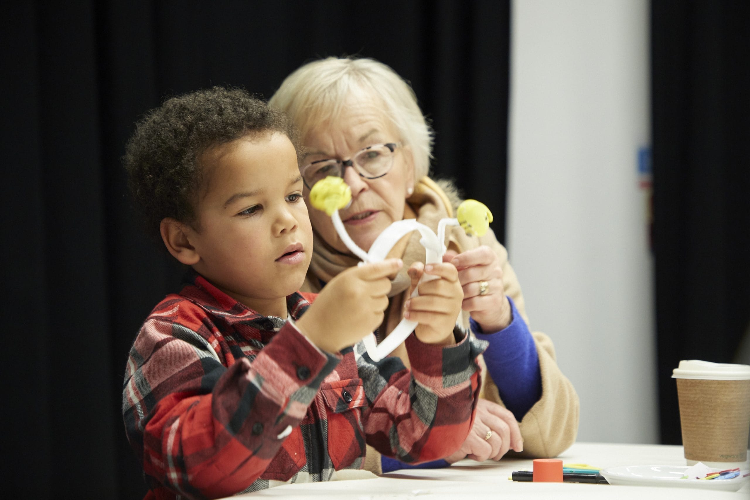 A small boy with dark hair and a checked shirt crafts with his grandma