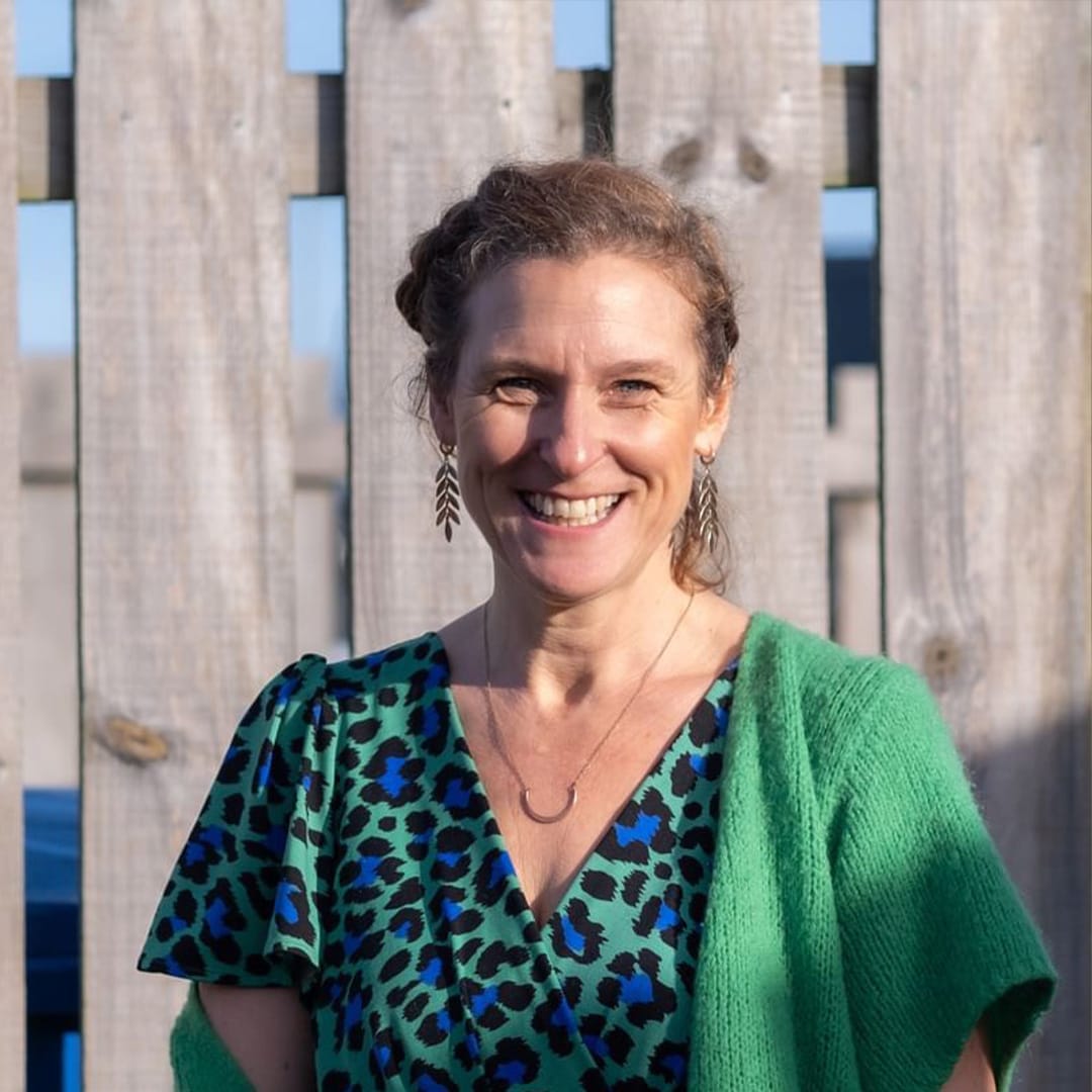 A lady wearing a green leopard print top stands in front of a fence.