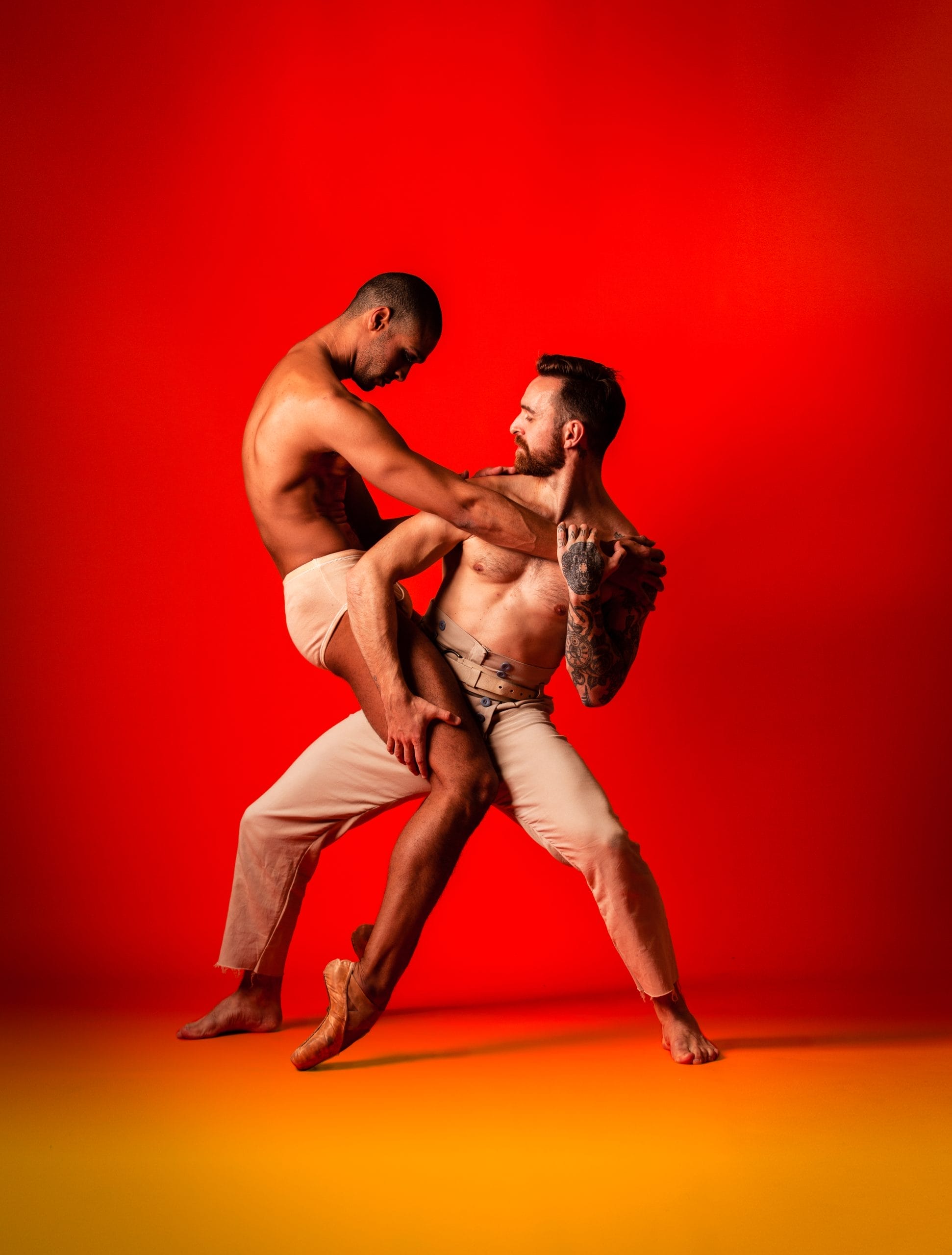 Two shirtless men dance together, with a red backdrop