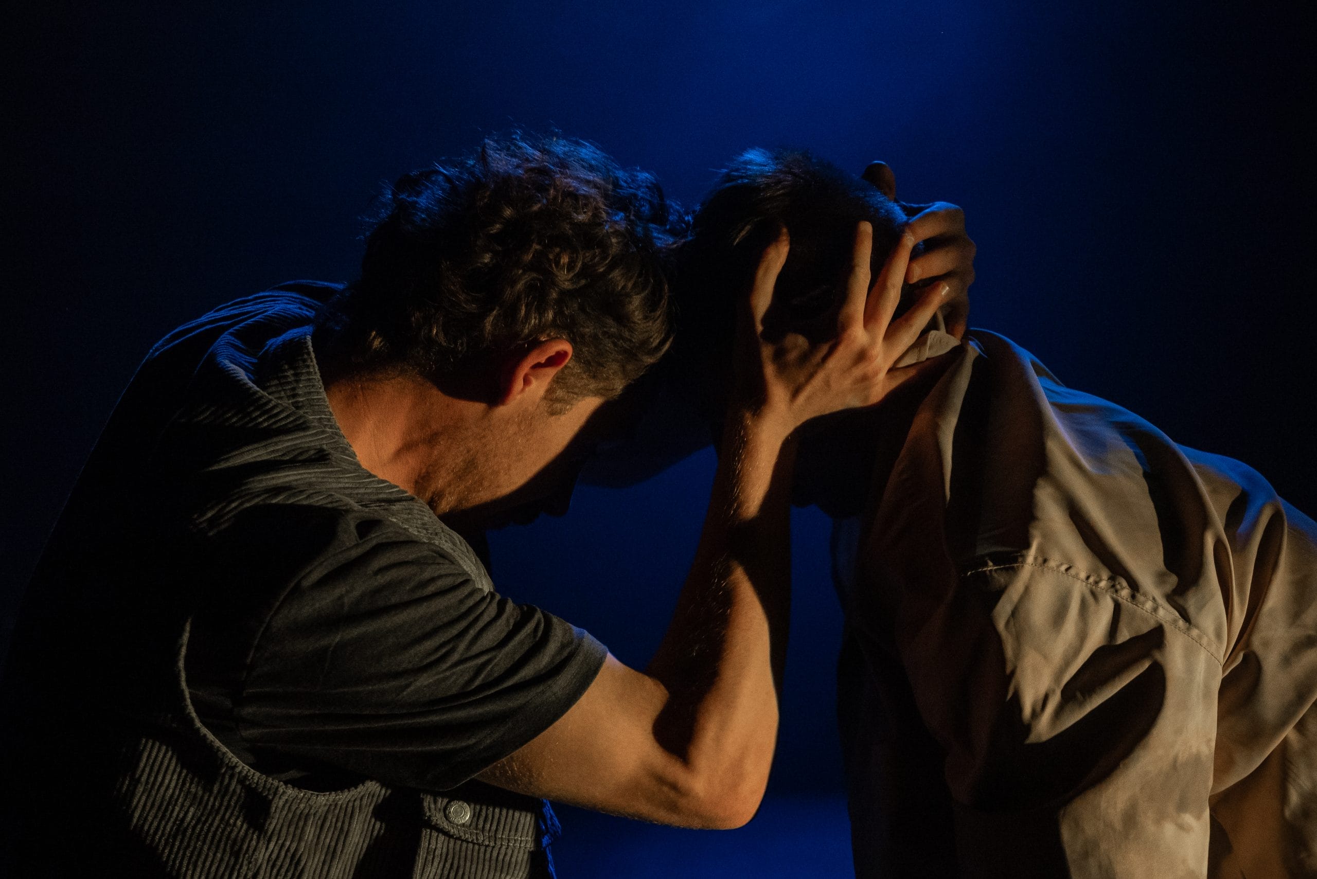 Two people perform on a dark stage. Only the back of their heads are visible and one holds the other's head in his hands.