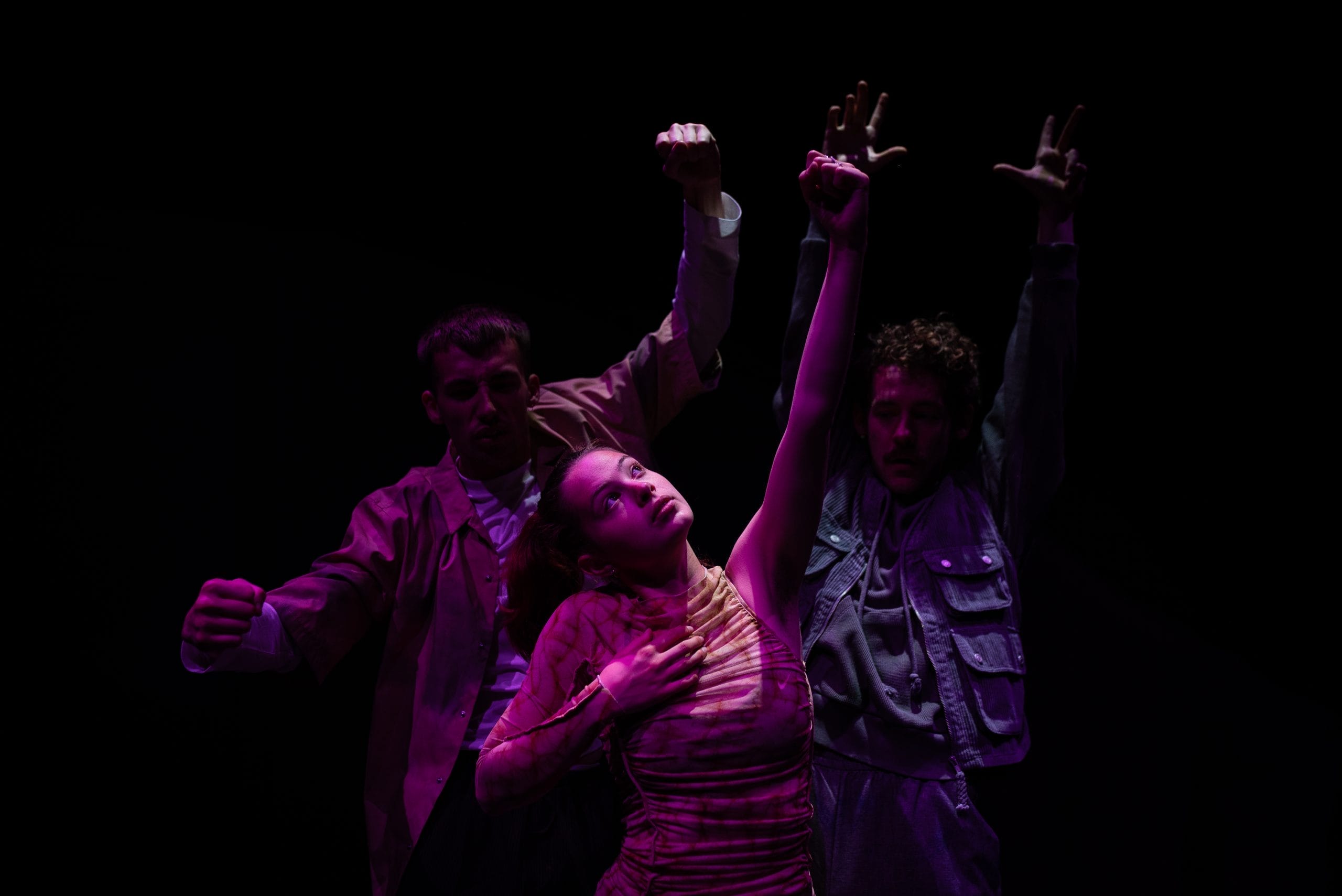 three performers dance on a dark stage. The woman in the middle looks up and they all reach up with a hand.