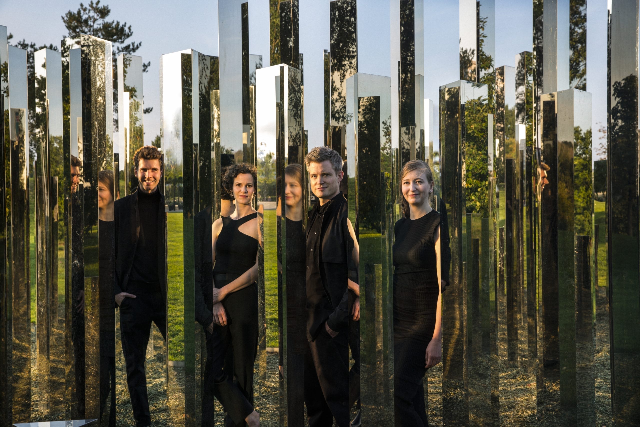 Two women and two men dressed in black standing in a grassy field in-between lots of fragments of mirrors