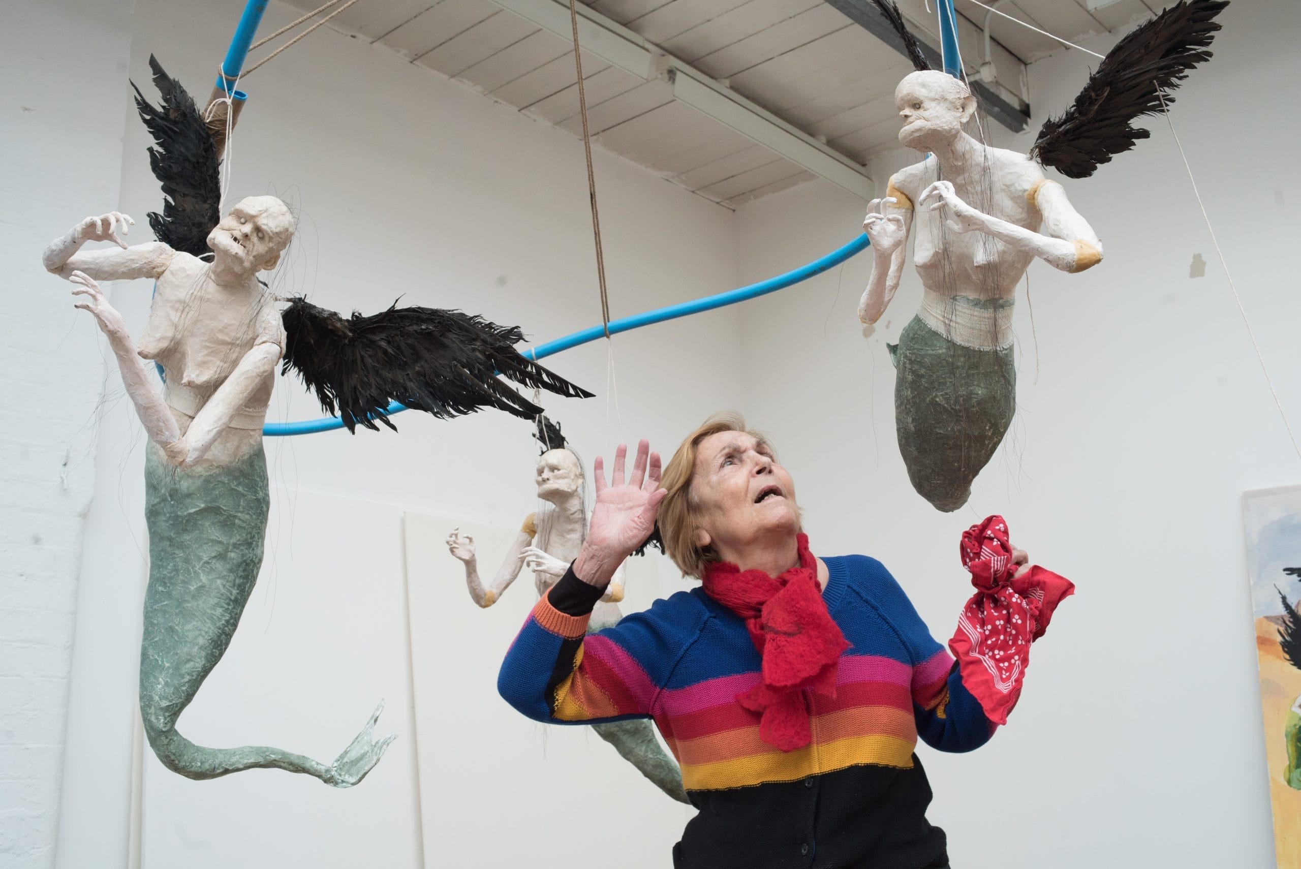 A woman, wearing a bright striped jumper and red scarf is surrounded by a swarm of flying mermaids.