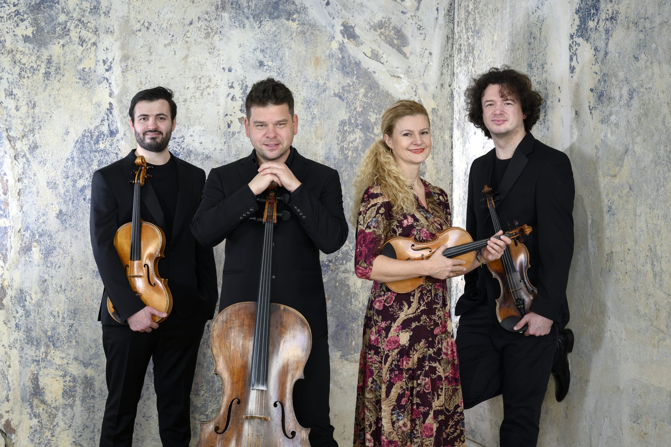 3 men dressed in black holding String instruments and one women in a floral dress standing in front of a greyish blue backdrop.