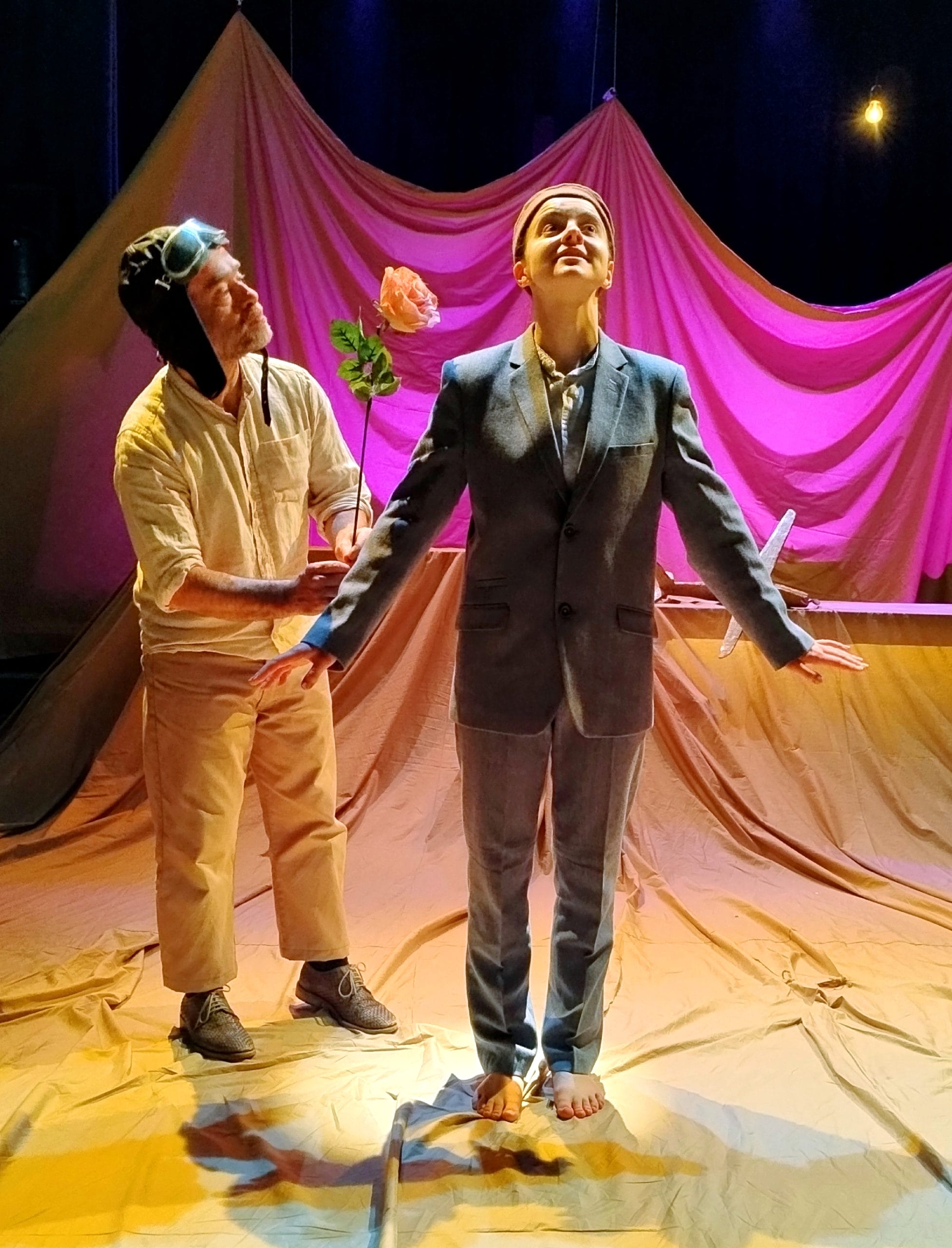 two actors stand on a stage, one dressed as a pilot offering a rose to the other who smiles
