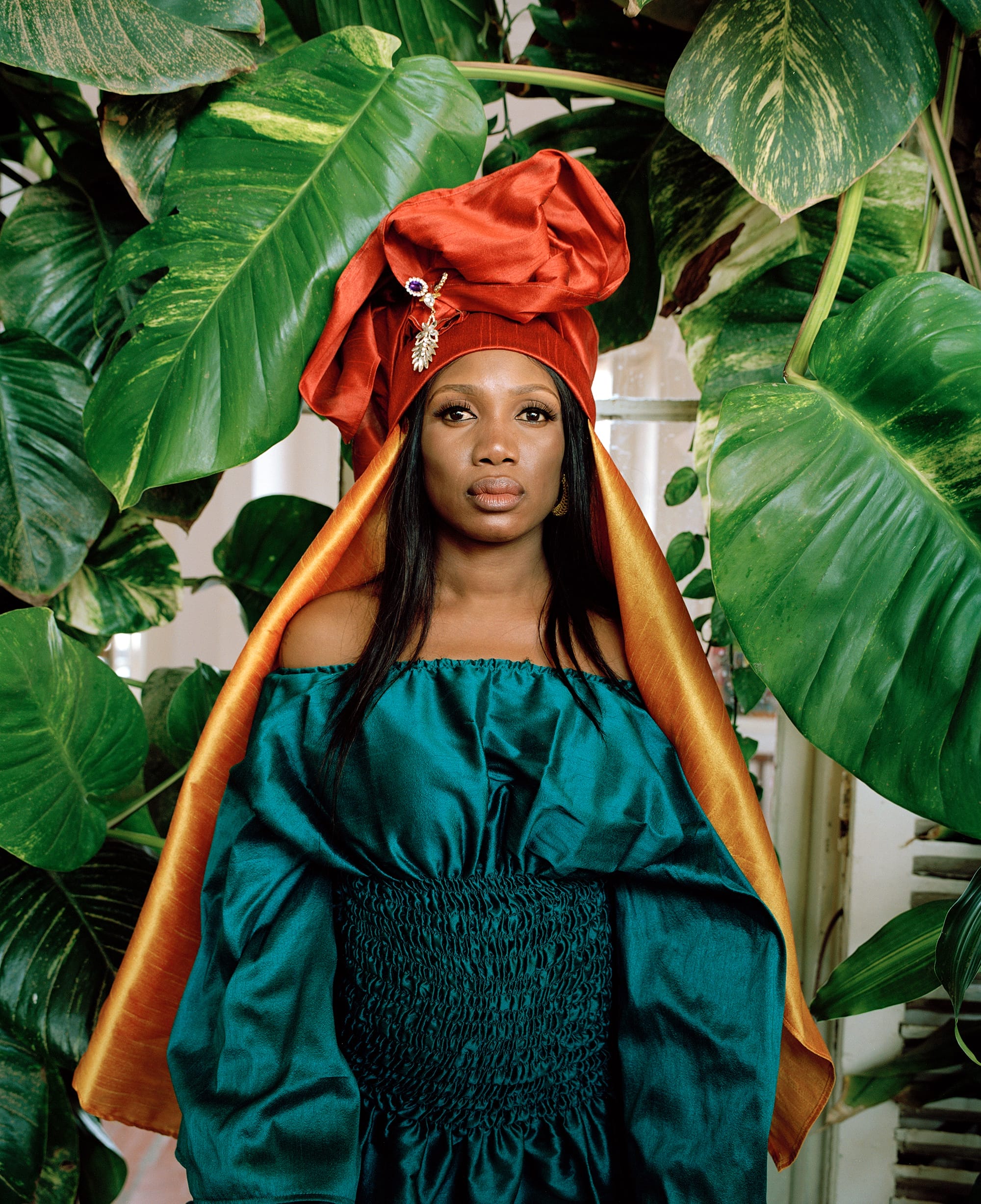 Woman wearing blue dress, red head wrap and golden veil standing in front of leafy backdrop.