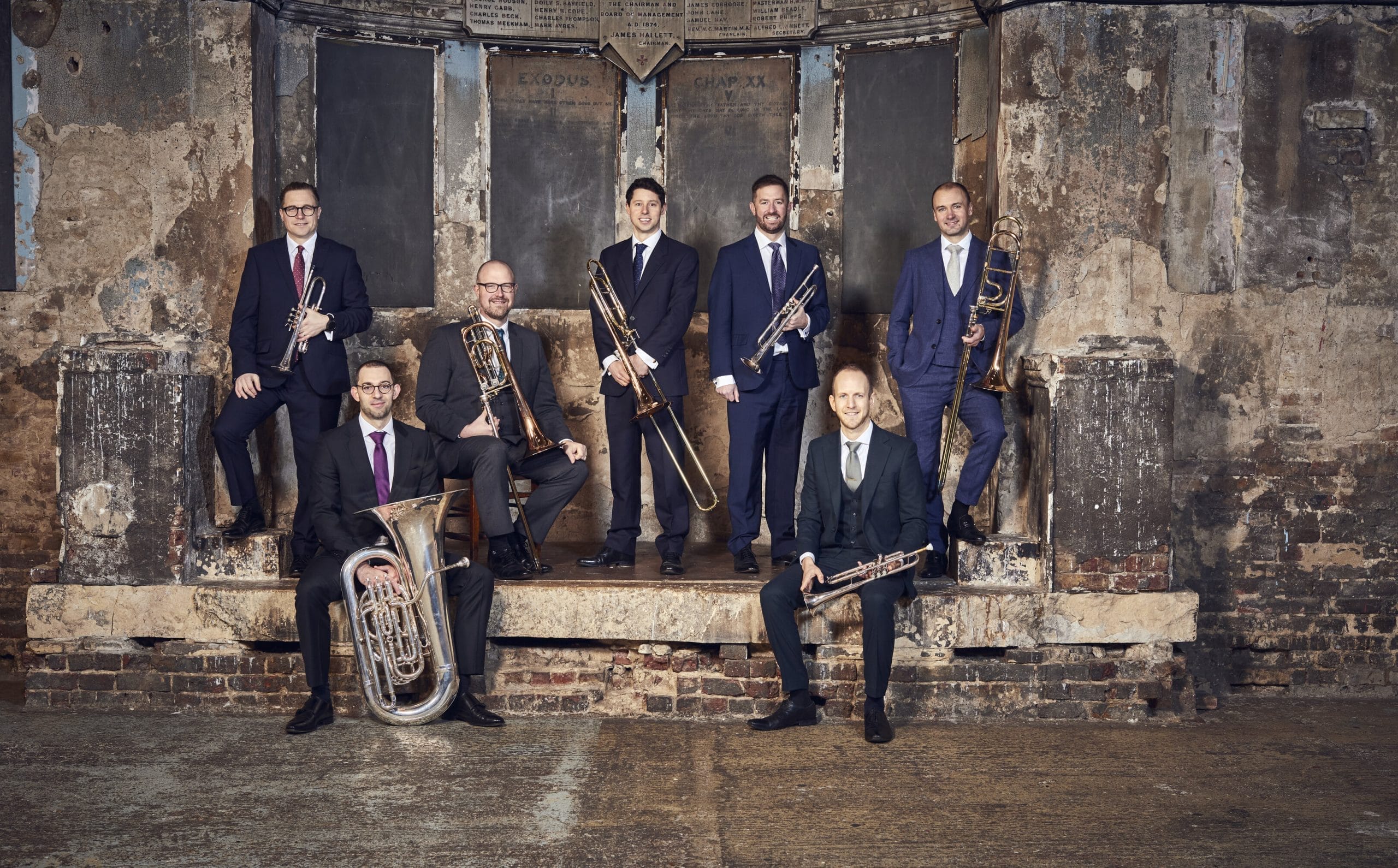 7 men dressed in dark suits with brass instruments stood in an industrial type set