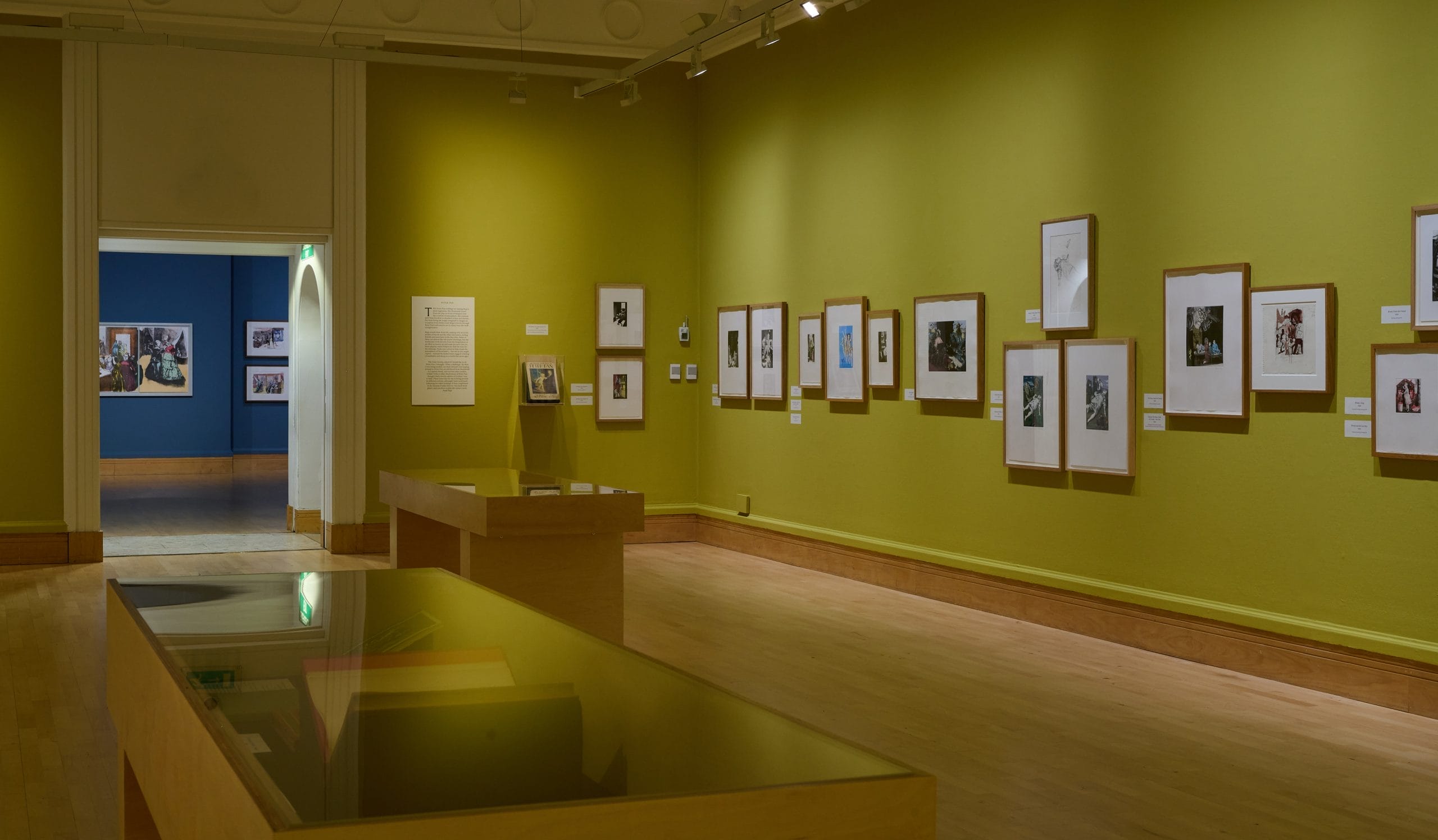 A gallery space with yellow walls, featuring a display case and prints by Paula Rego on the wall.